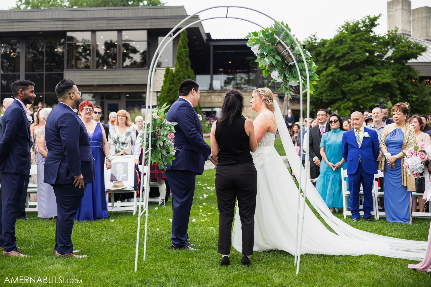 Wedding ceremony under white arch with guests in blue and formal attire standing on lawn.