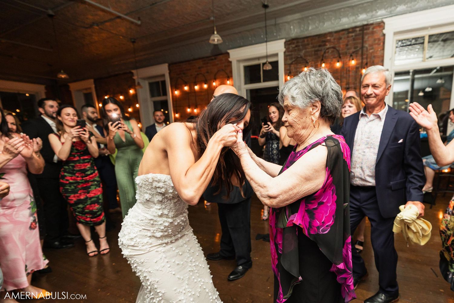Wedding guests dance and celebrate on a rustic indoor dance floor with string lighting.
