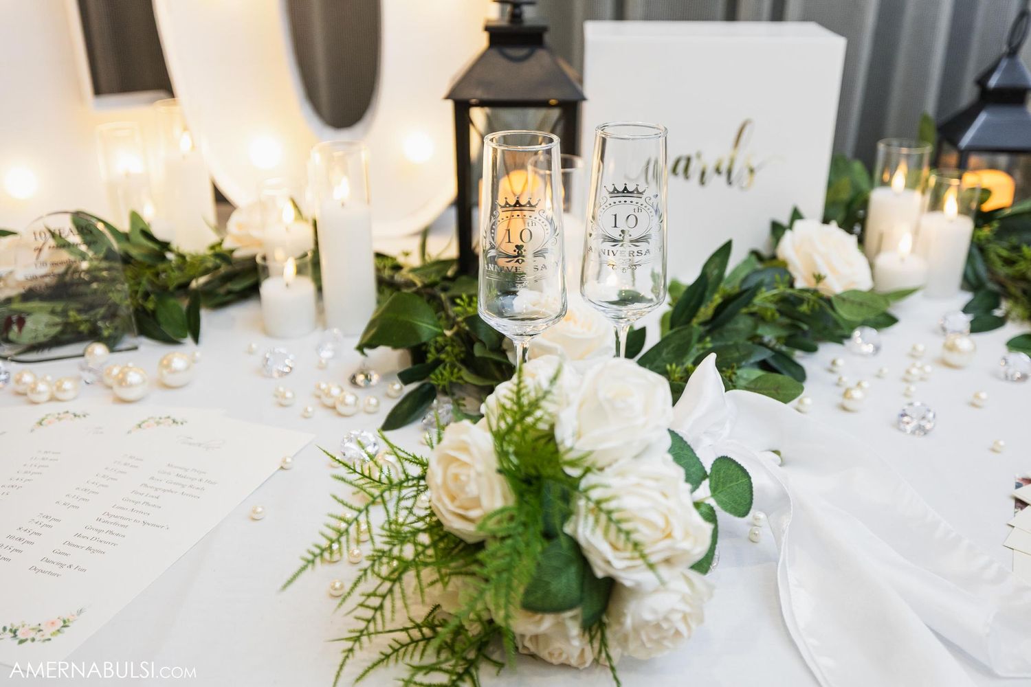 Elegant wedding table setting with white roses, greenery, and lantern centerpieces.