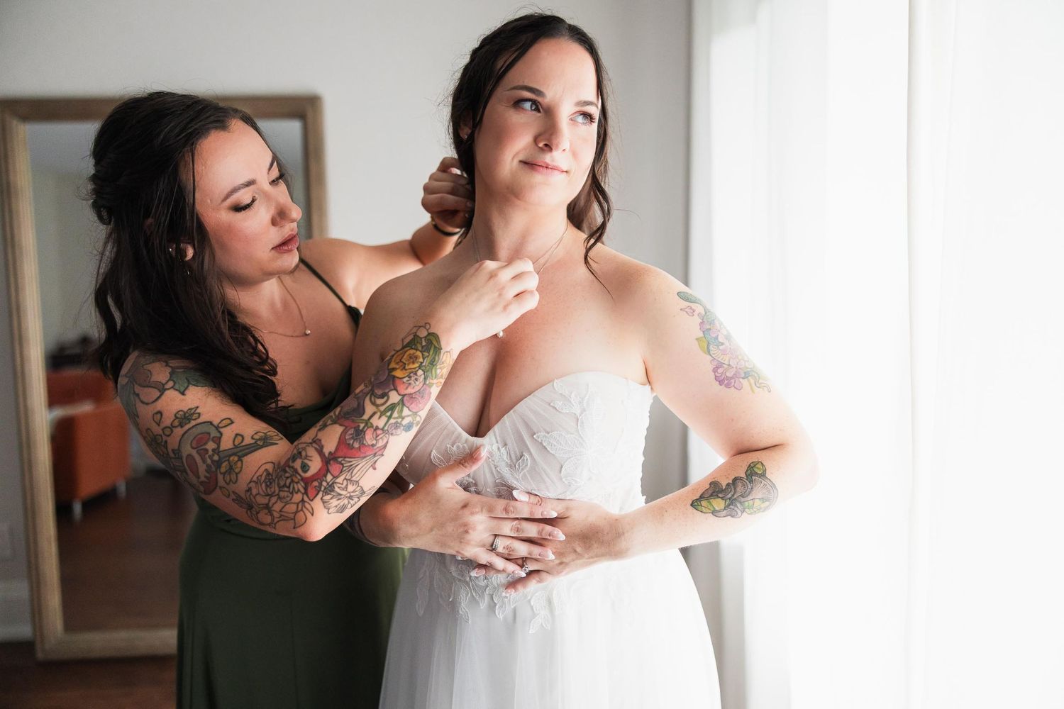 A bridesmaid helps adjust the bride's dress while getting ready.