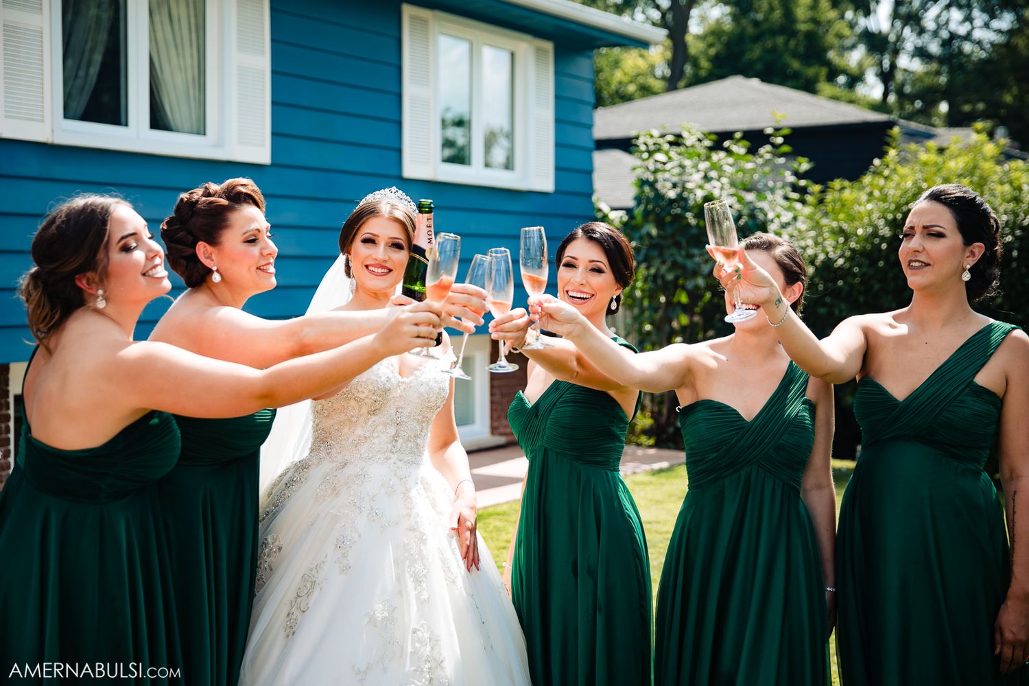Wedding party in emerald green dresses toasting with champagne glasses outside blue house.