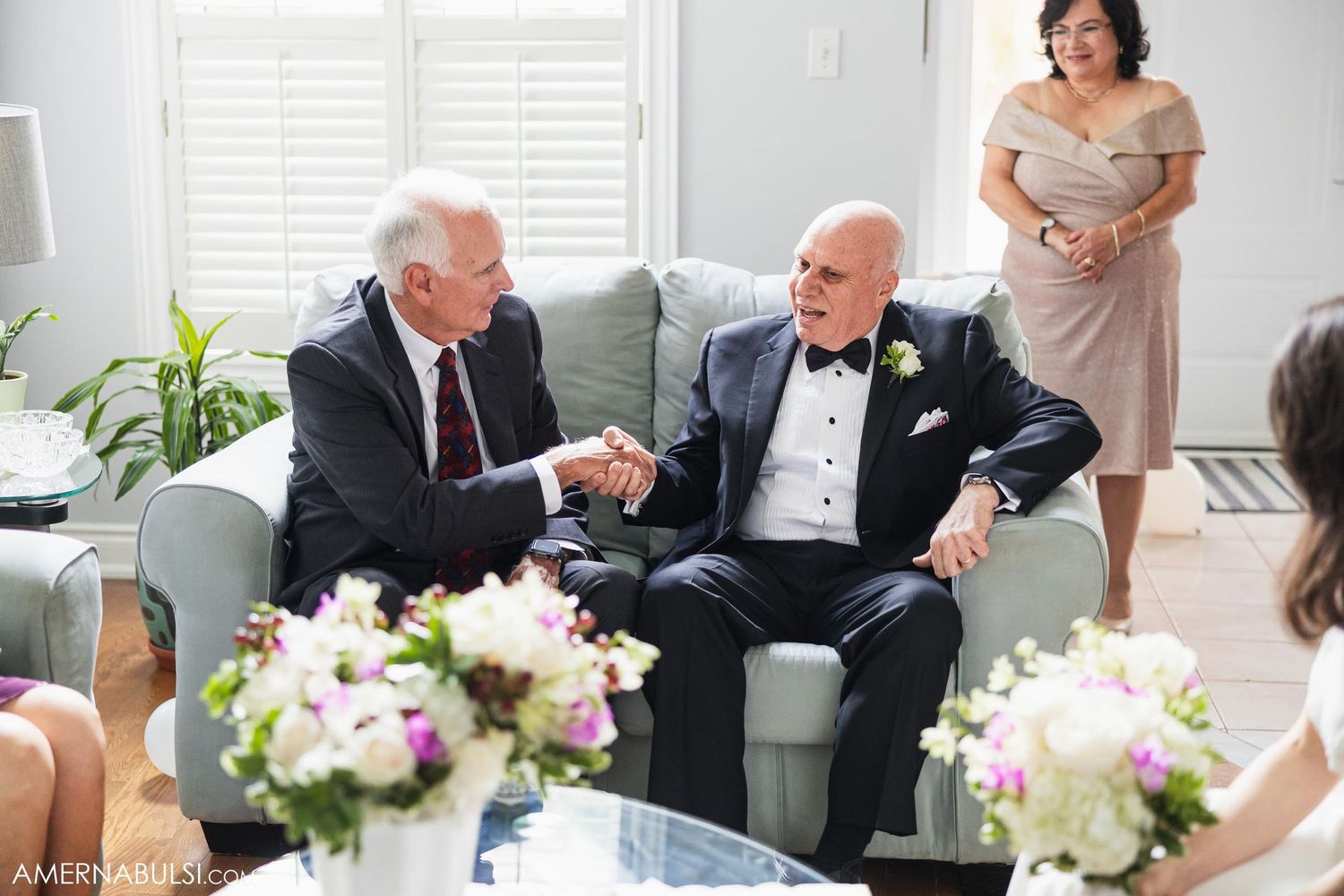 Two people share a conversation on a couch while elegant floral arrangements decorate the room at a formal event.