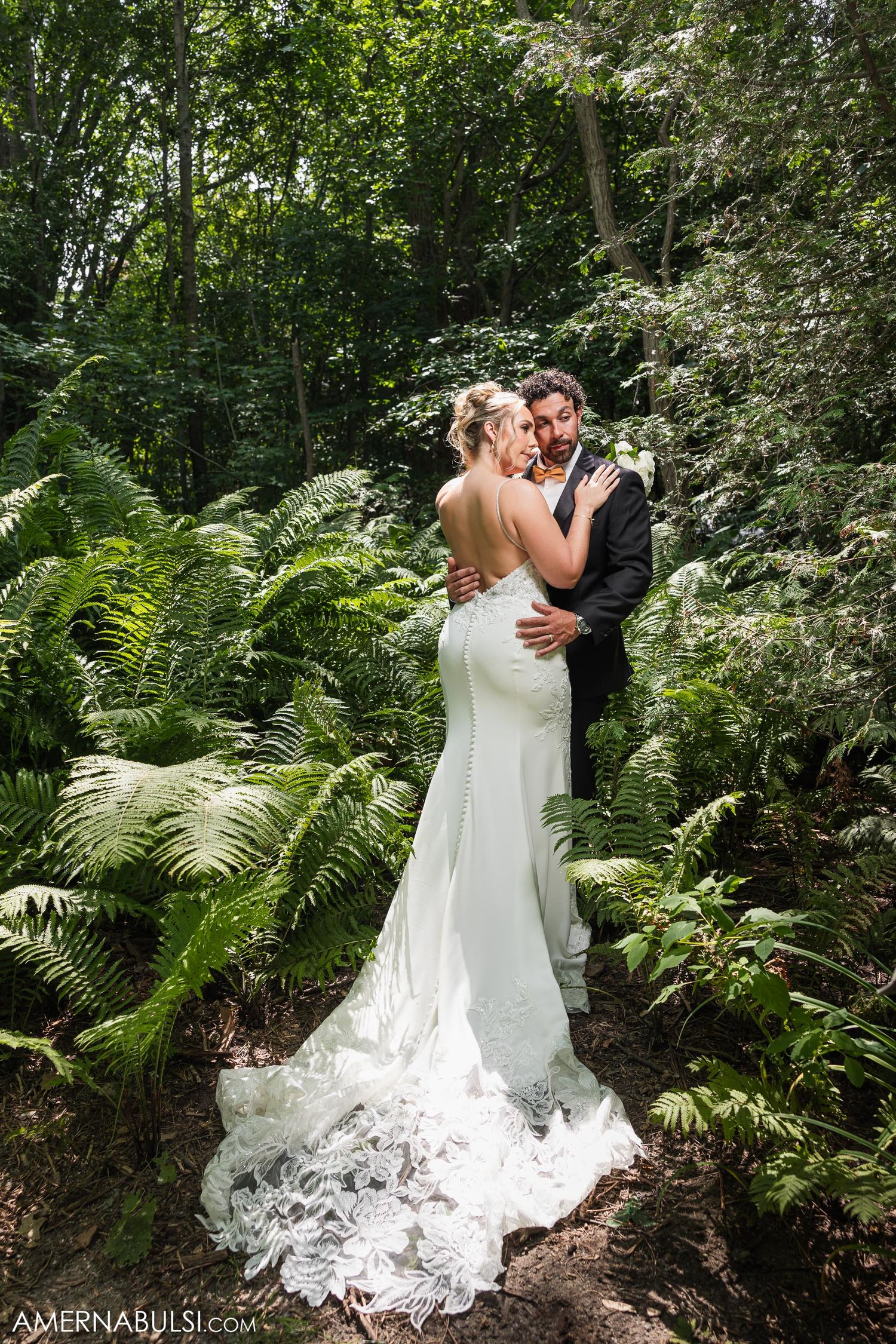 Couple shares romantic moment in forest surrounded by ferns, bride wearing elegant fitted gown with lace train.