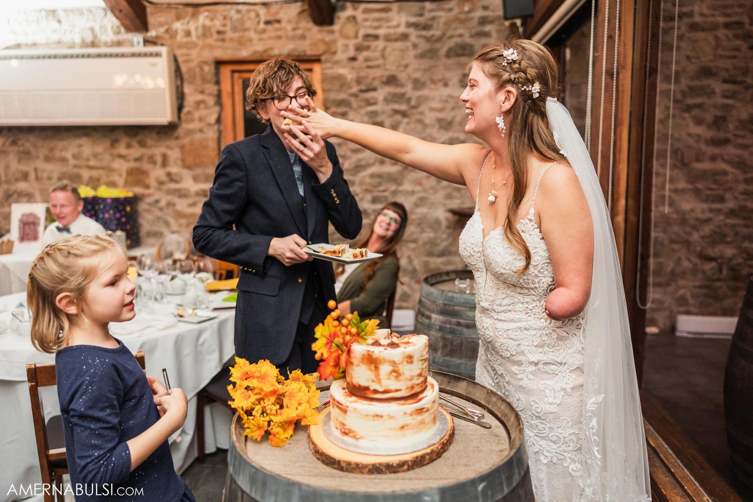 Fun cake-feeding moment during wedding reception with young onlooker watching.