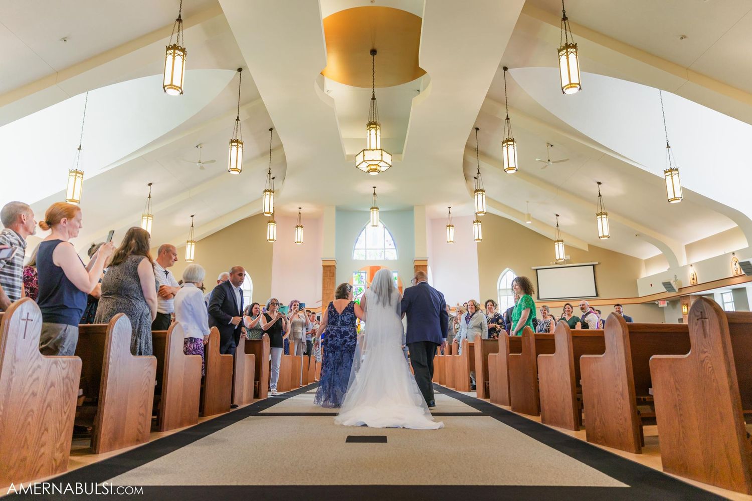 A wide-angle view of a church wedding ceremony with guests seated in wooden pews under hanging lights.