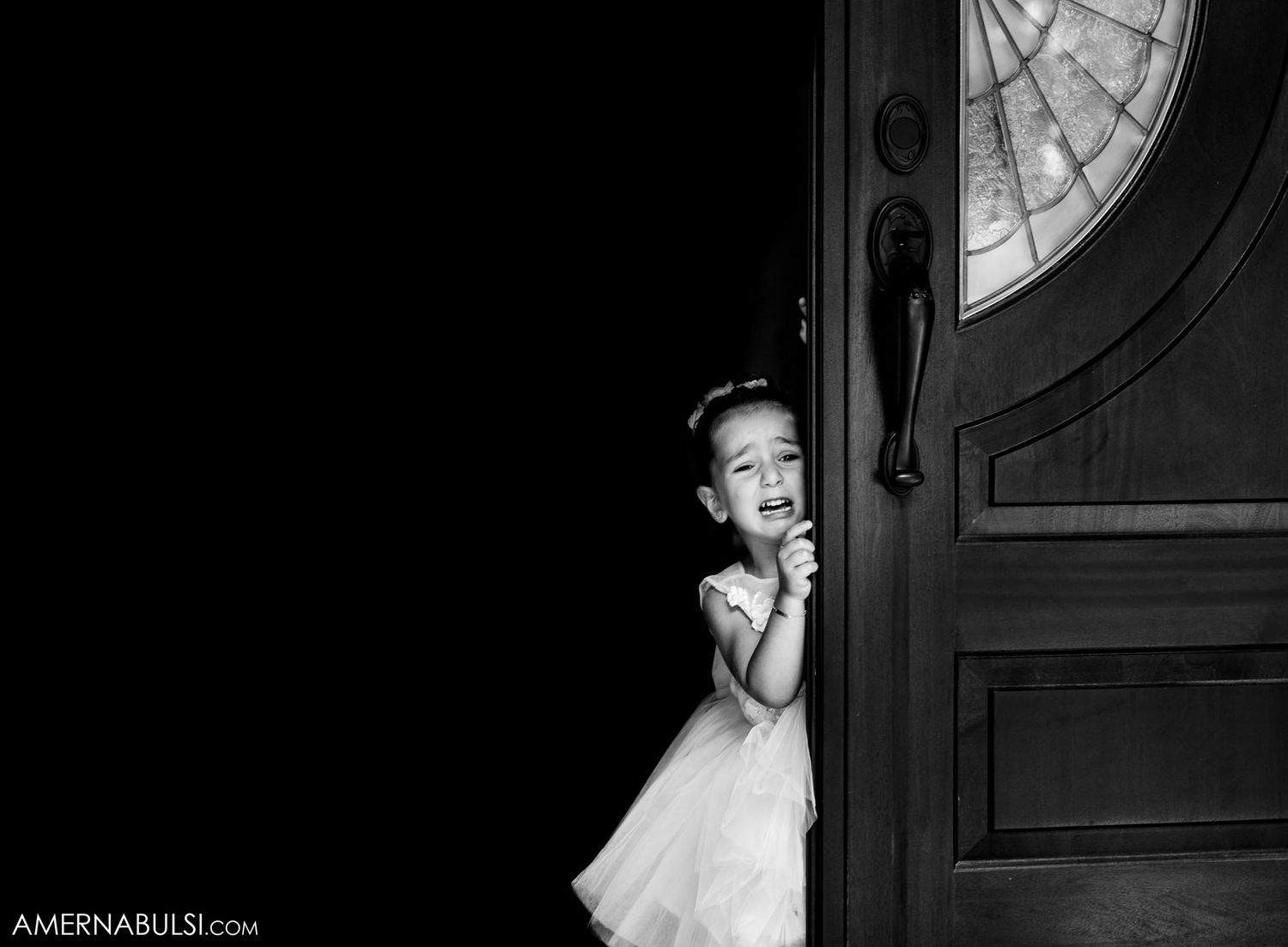 Artistic black and white image of a small child in white dress peeking around an elegant doorway.