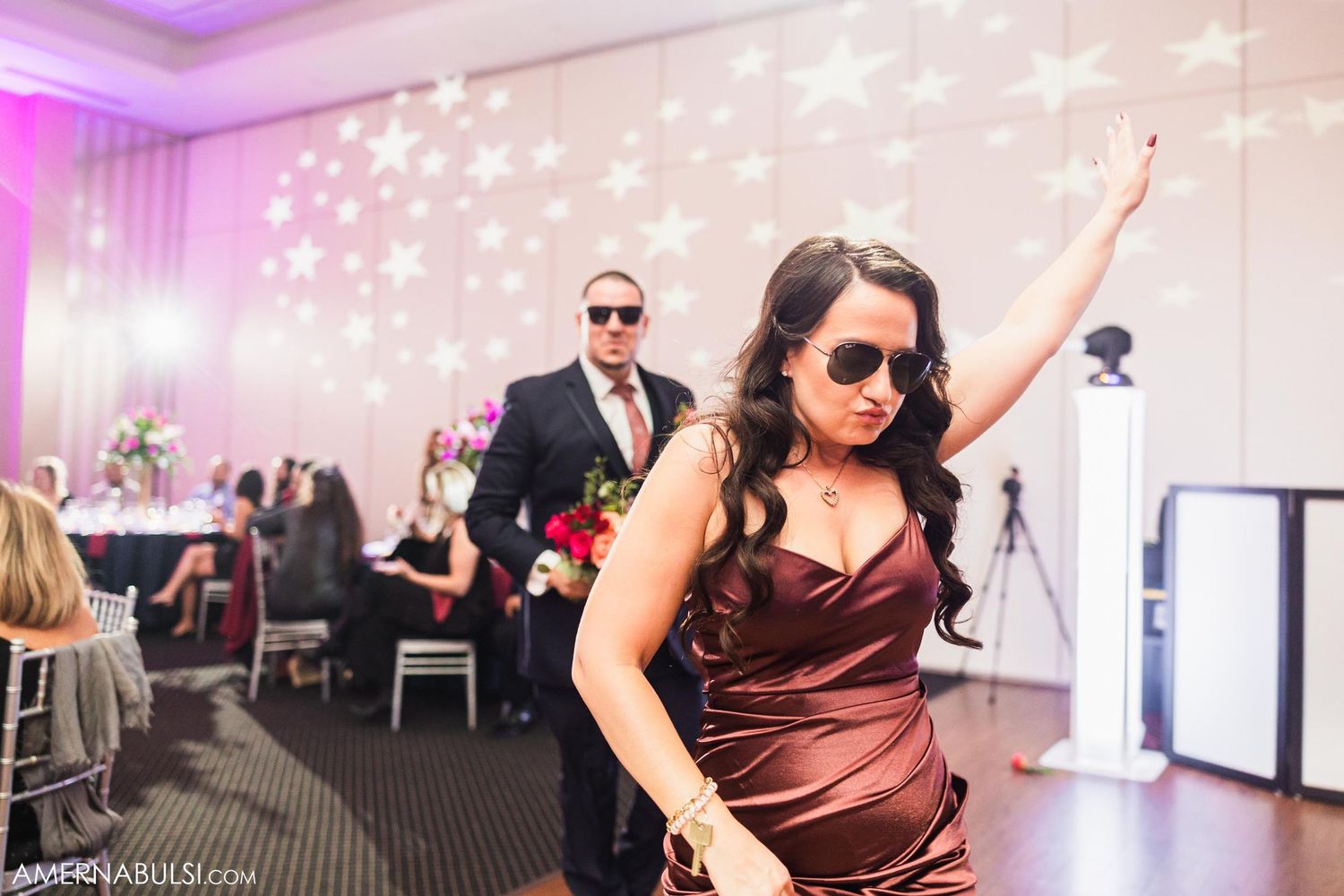 Wedding guest in satin burgundy dress dances at reception with pink uplighting and starry backdrop.