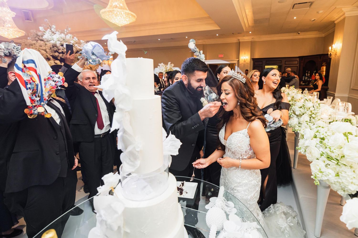 Wedding celebration around ornate white tiered cake with guests gathered in elegant ballroom setting.