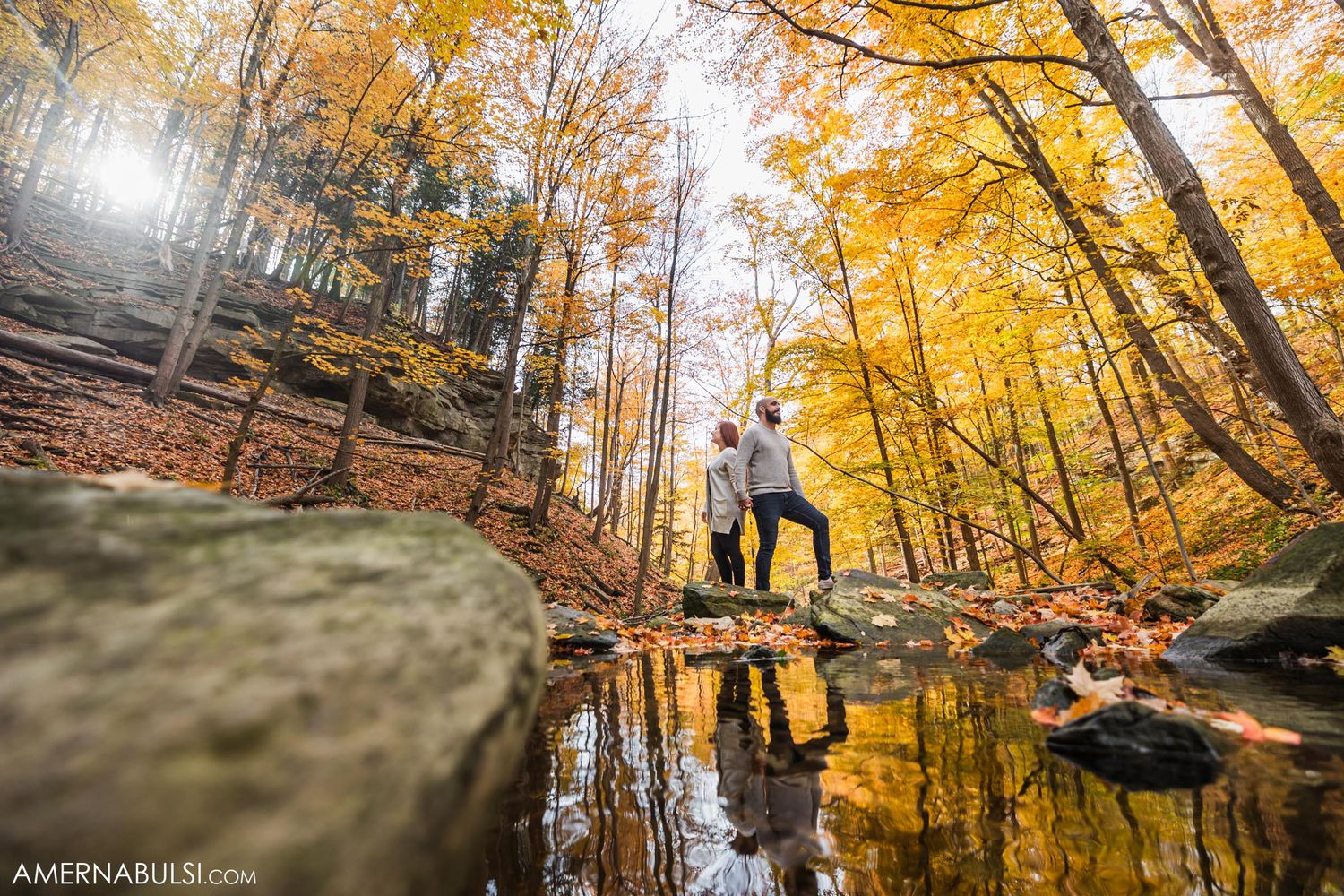 Romantic couple poses by reflective pond surrounded by golden autumn trees.