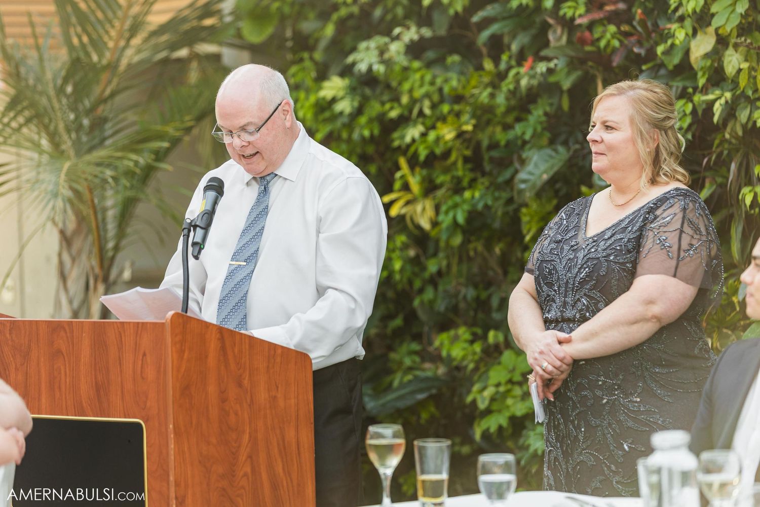 A speaker delivers remarks at an outdoor wedding podium with lush greenery in the background.