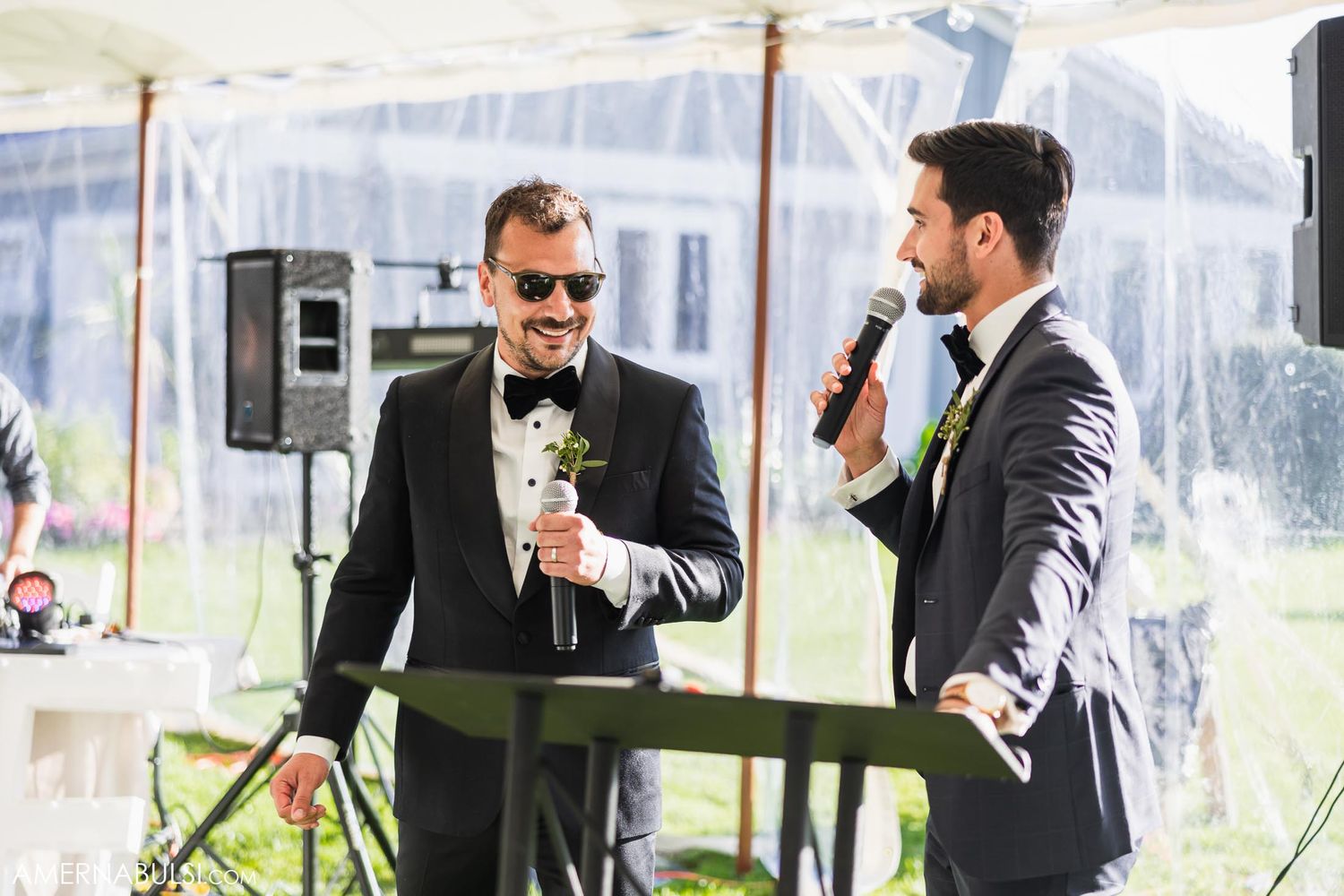 Two men in formal attire speaking into microphones at outdoor wedding reception.