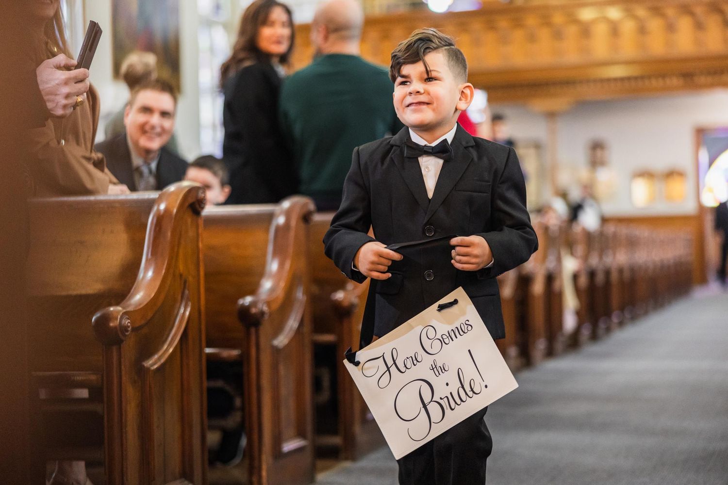 Young boy in formal attire carries wedding sign while walking down church aisle.