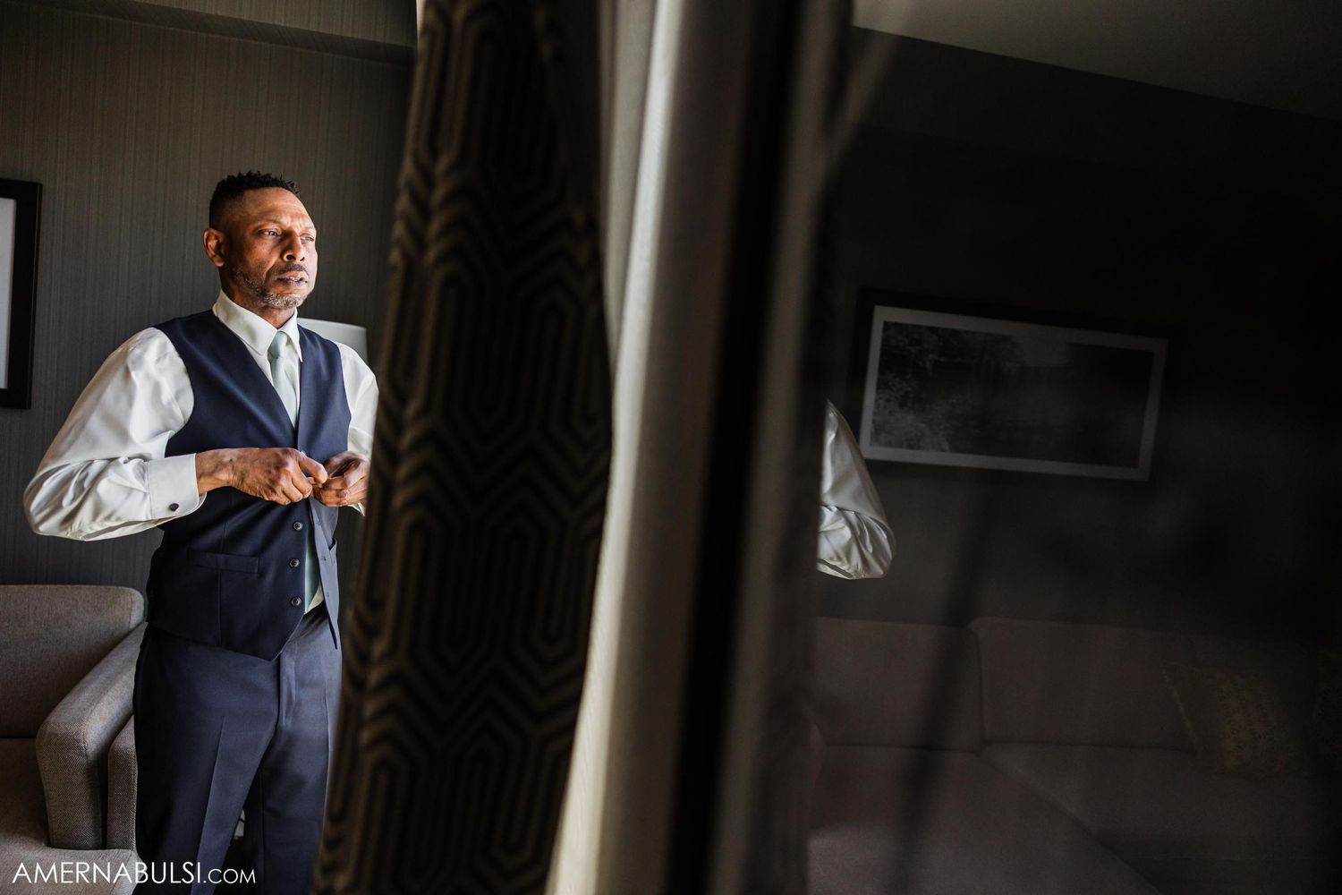 A groom in a navy vest and white shirt adjusts his cufflinks while getting ready in a hotel room.