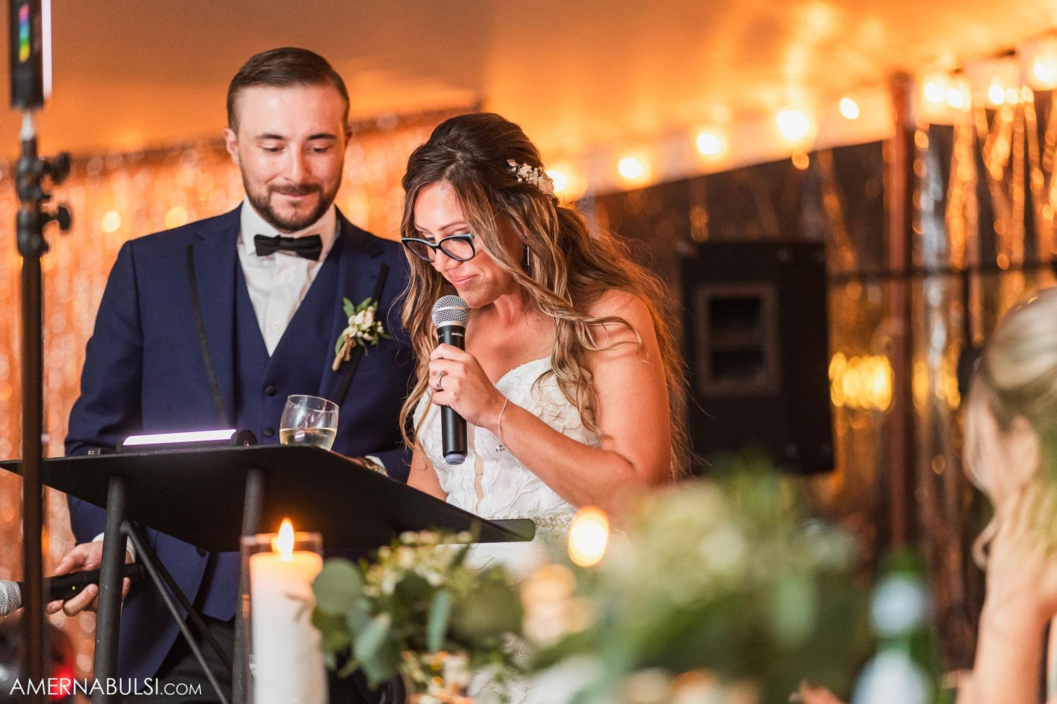 Couple delivering heartfelt wedding speeches at an outdoor reception under glowing string lights.