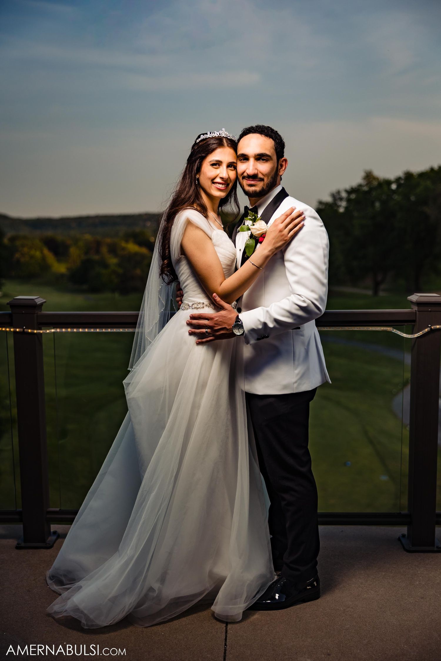 A couple in wedding attire pose together on an outdoor balcony with scenic views.