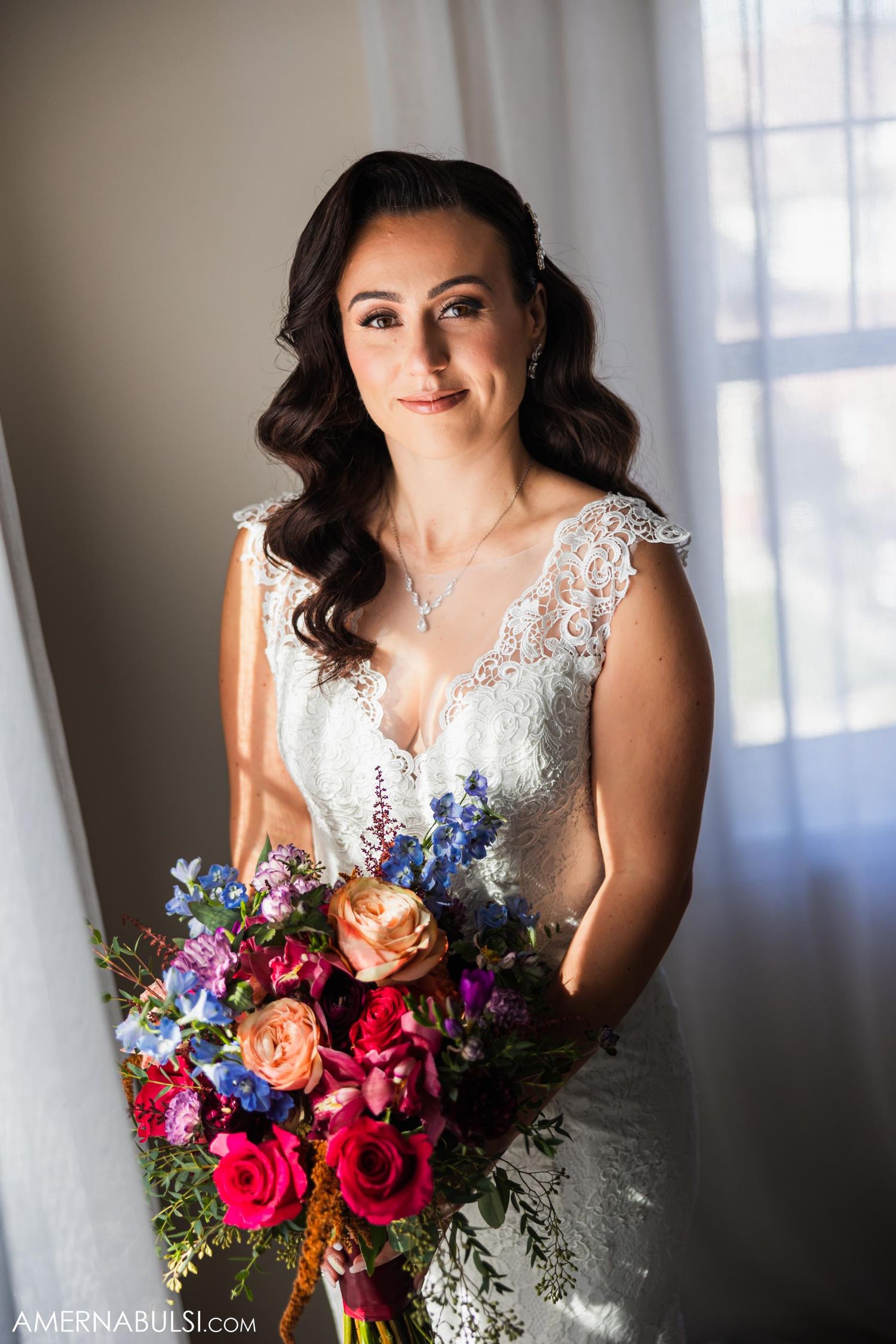 A bride poses by a window in a lace wedding gown holding a colorful bouquet.