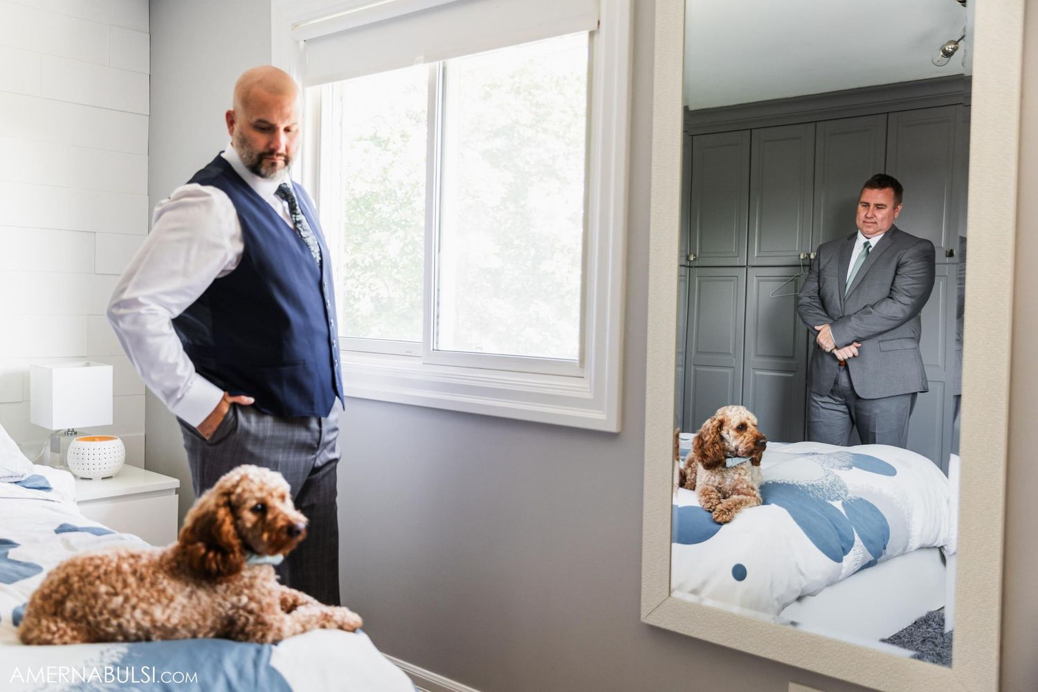 A groom in a blue vest stands in a bedroom with a brown dog while looking in a mirror.