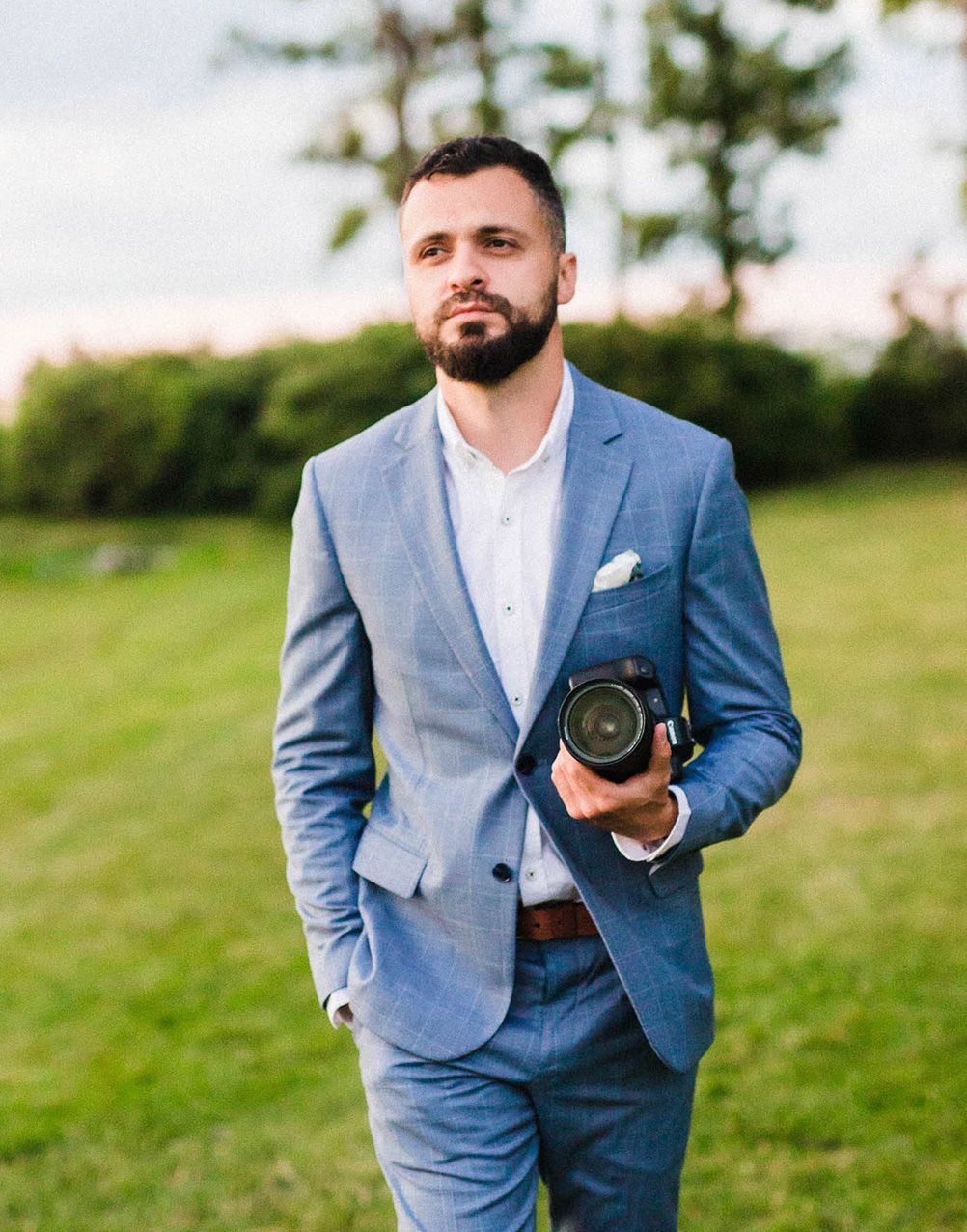 Wedding photographer in light blue suit holds professional camera on grassy hill.