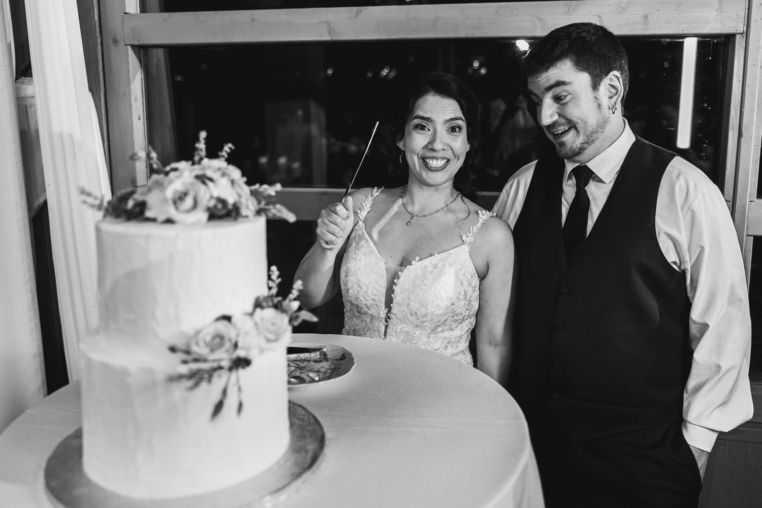 Bride and groom smile while cutting a two-tier white wedding cake decorated with flowers.