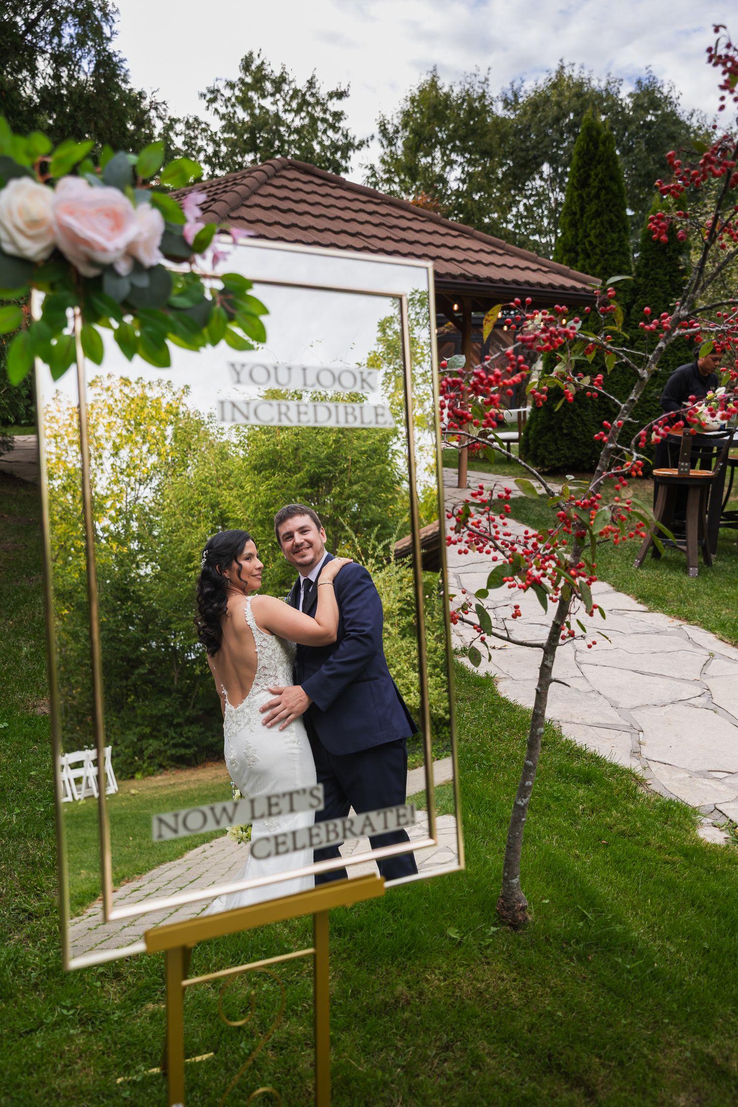 Bride and groom posing inside a decorative gold mirror frame sign reading Now Lets Celebrate outdoors.