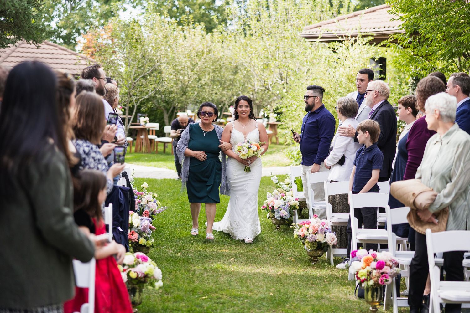 Bride walking down the aisle in a white gown with her escort at a sunny outdoor garden wedding ceremony.