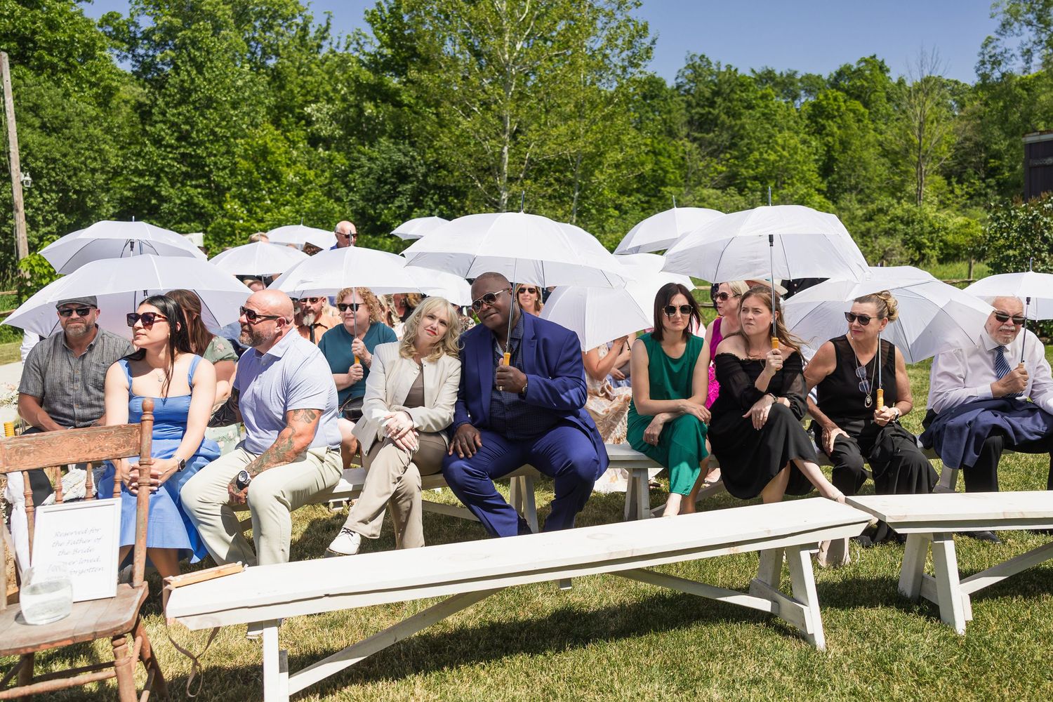 Wedding guests seated on white benches holding white parasol umbrellas during an outdoor ceremony.