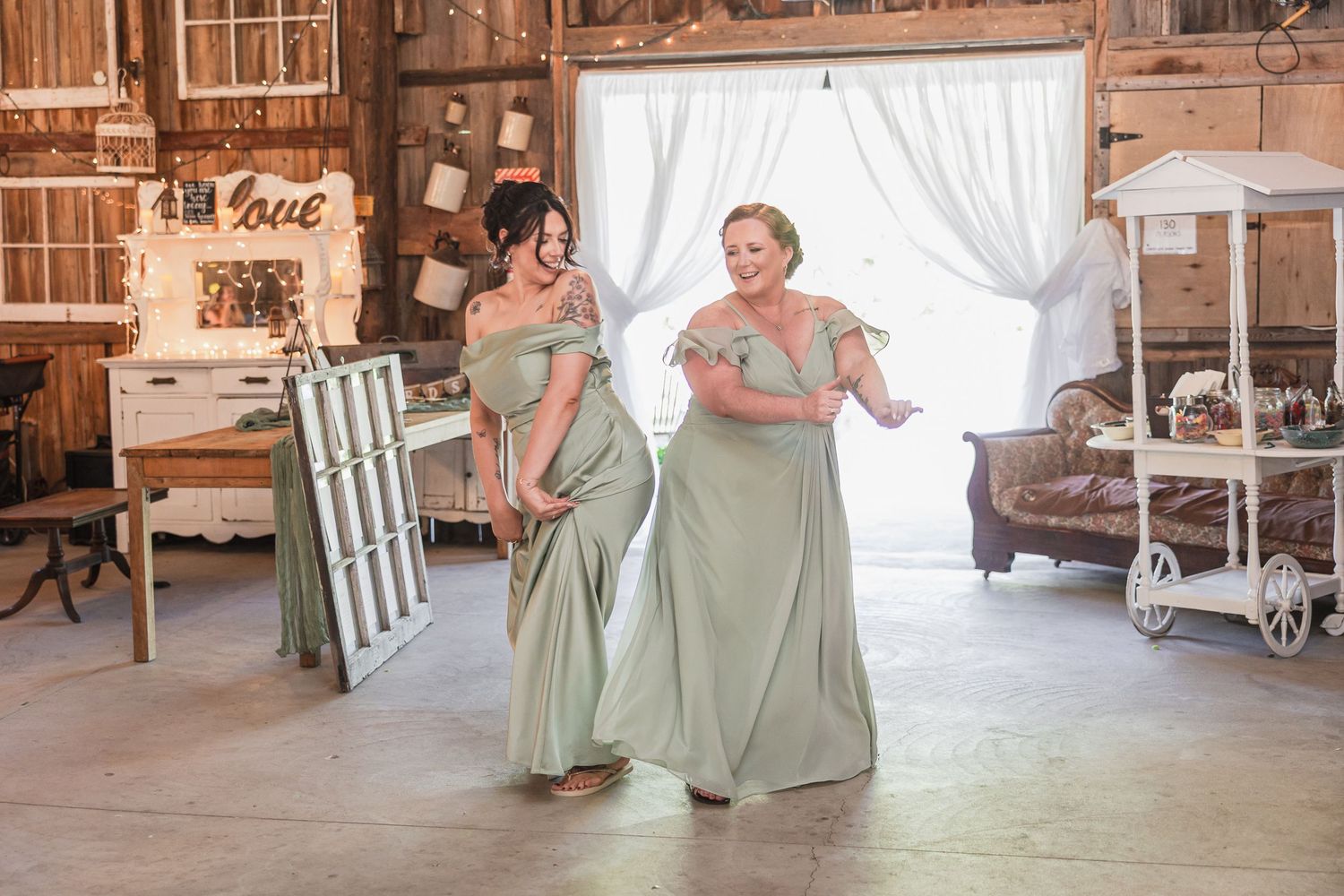 Two bridesmaids in sage green off-shoulder gowns dancing joyfully inside a rustic barn wedding venue.