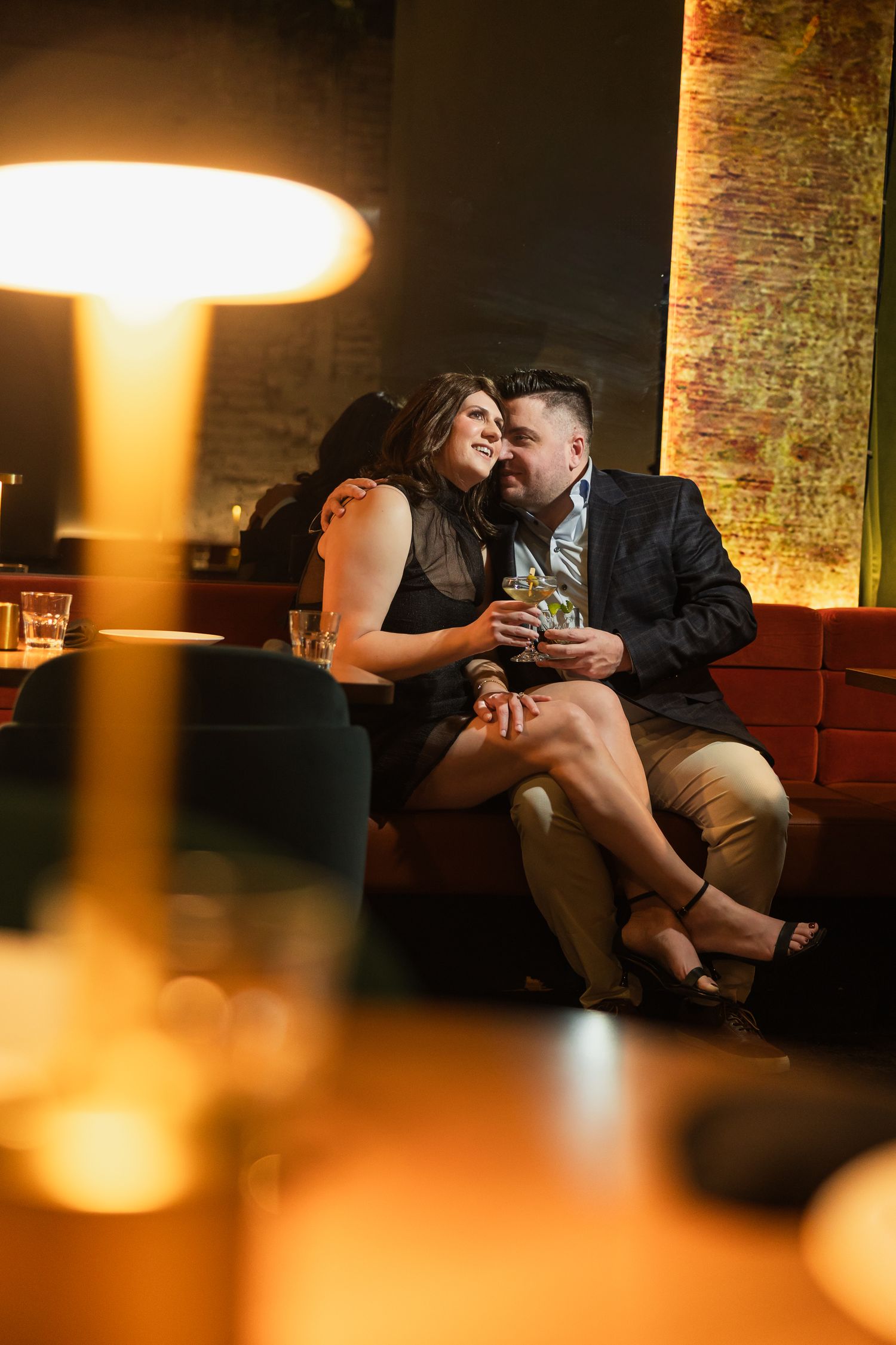 Couple sharing a romantic moment on a red velvet sofa in a dimly lit upscale bar with warm ambient lighting.