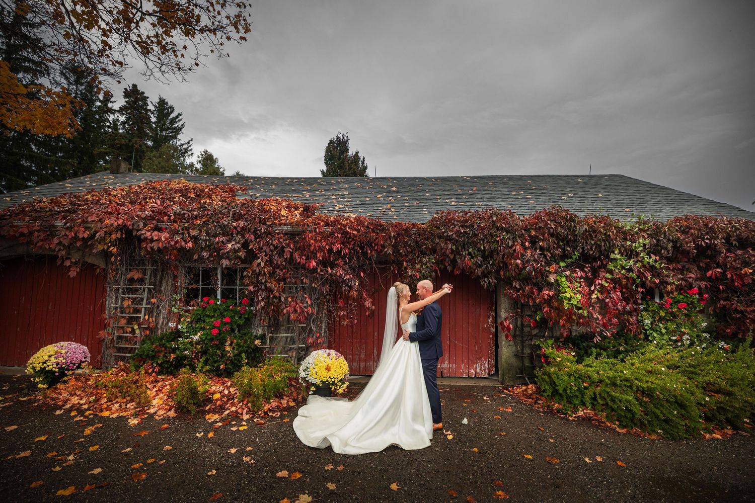 Wedding couple poses near red barn with autumn foliage and fallen leaves.
