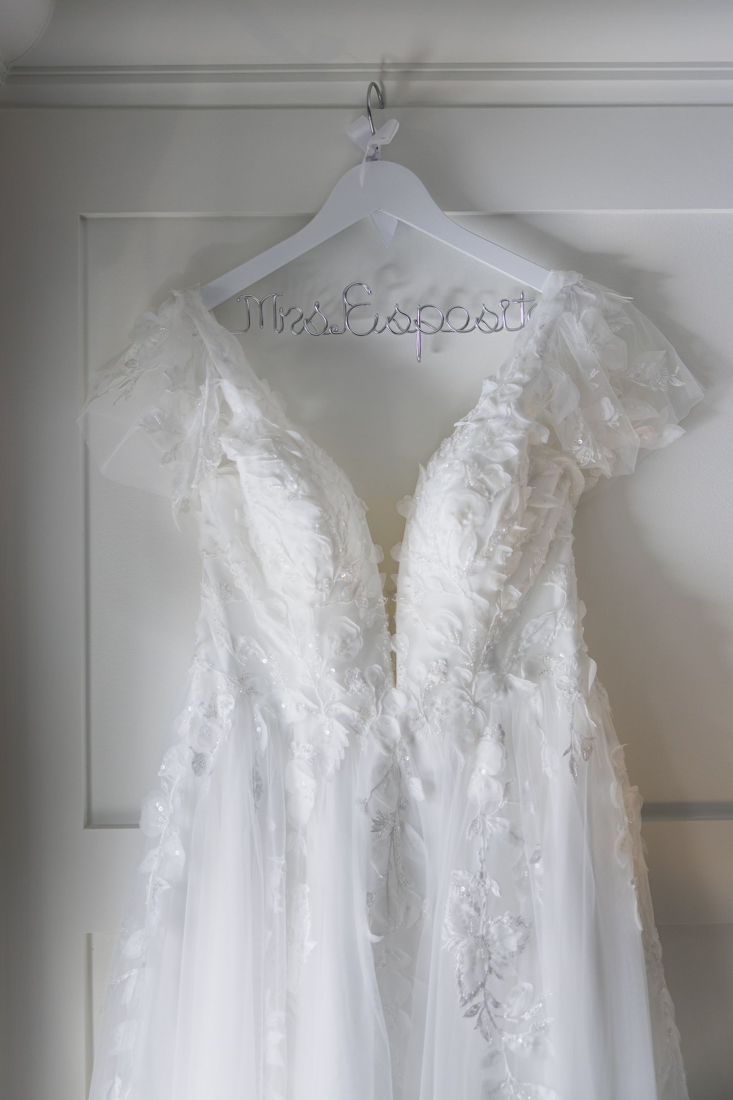 White lace A-line wedding dress hanging on personalized wire hanger reading Mrs. on white door.