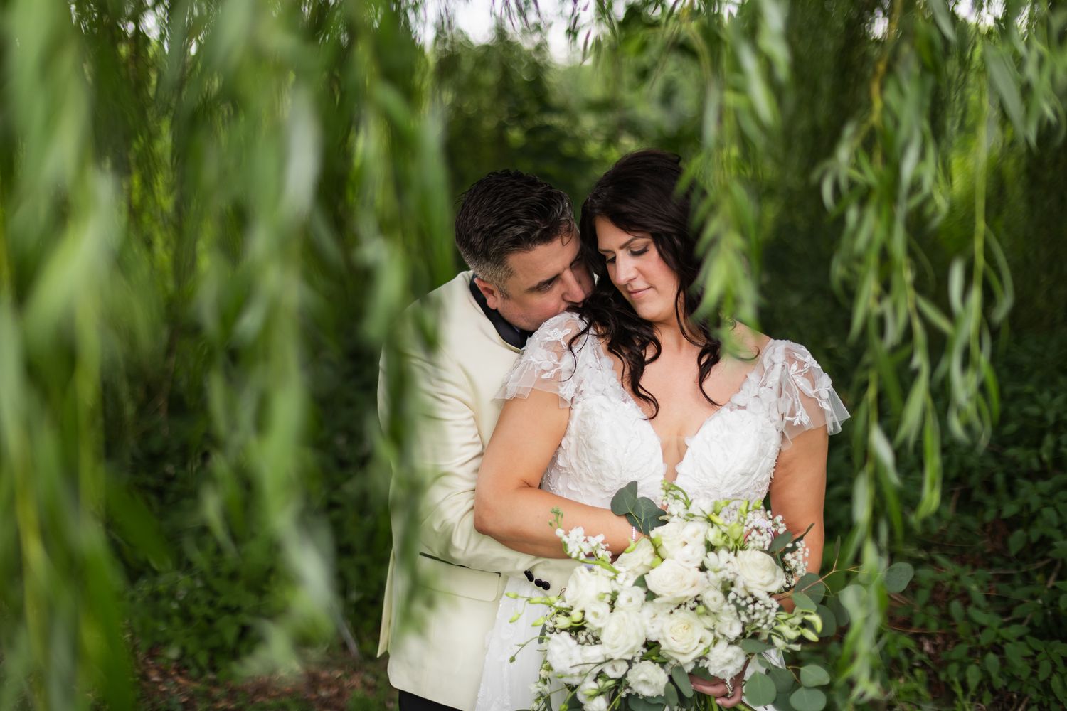 Bride and groom share a tender moment under weeping willow branches, bride holding white floral bouquet.