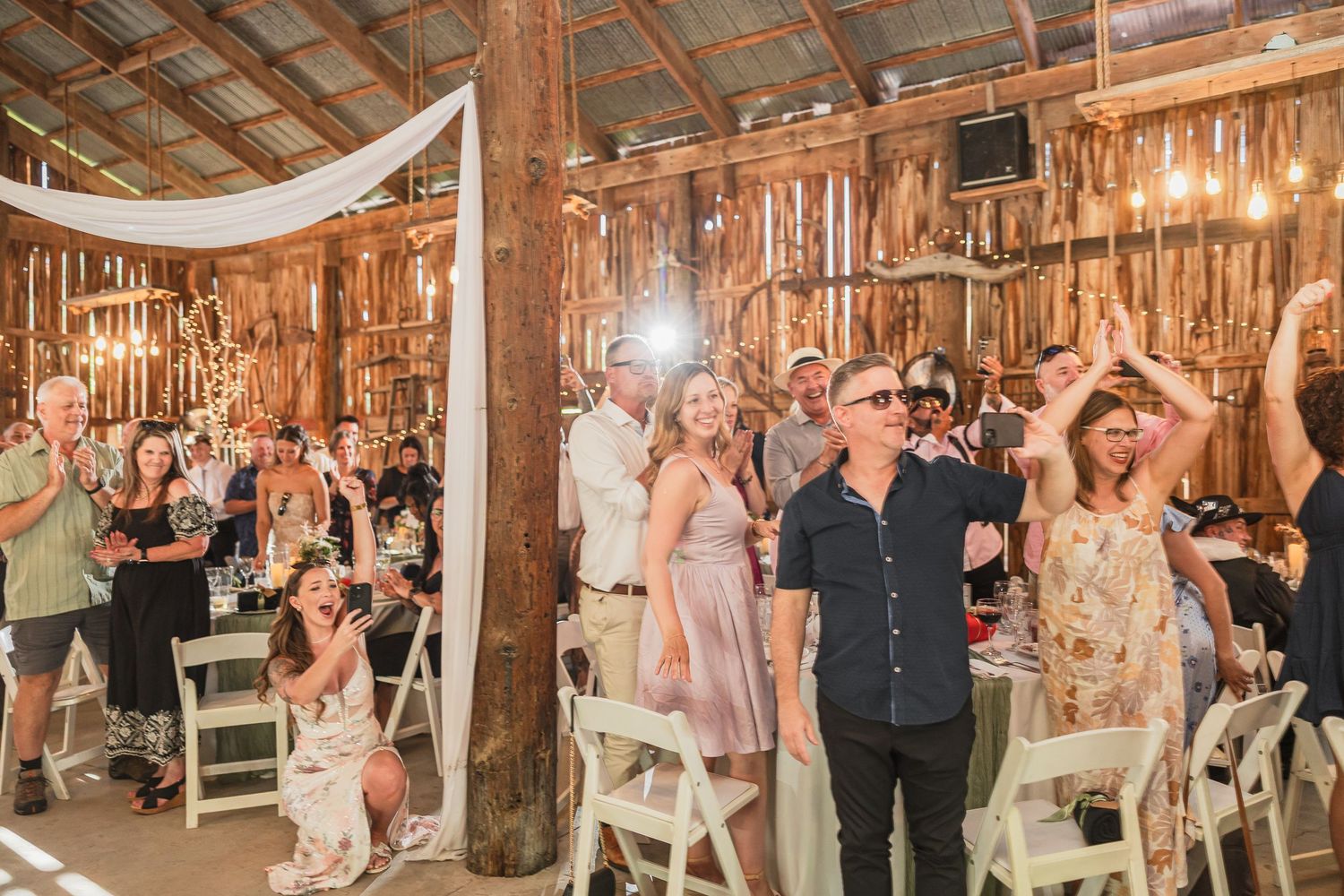 Guests dancing and celebrating at a rustic barn wedding reception with string lights and white chairs.