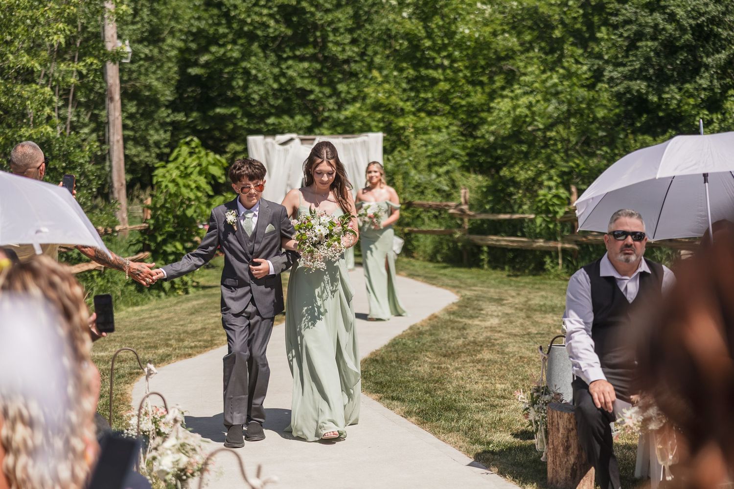 Bride in sage green gown walks down outdoor aisle with young groomsman as guests celebrate at summer wedding ceremony.