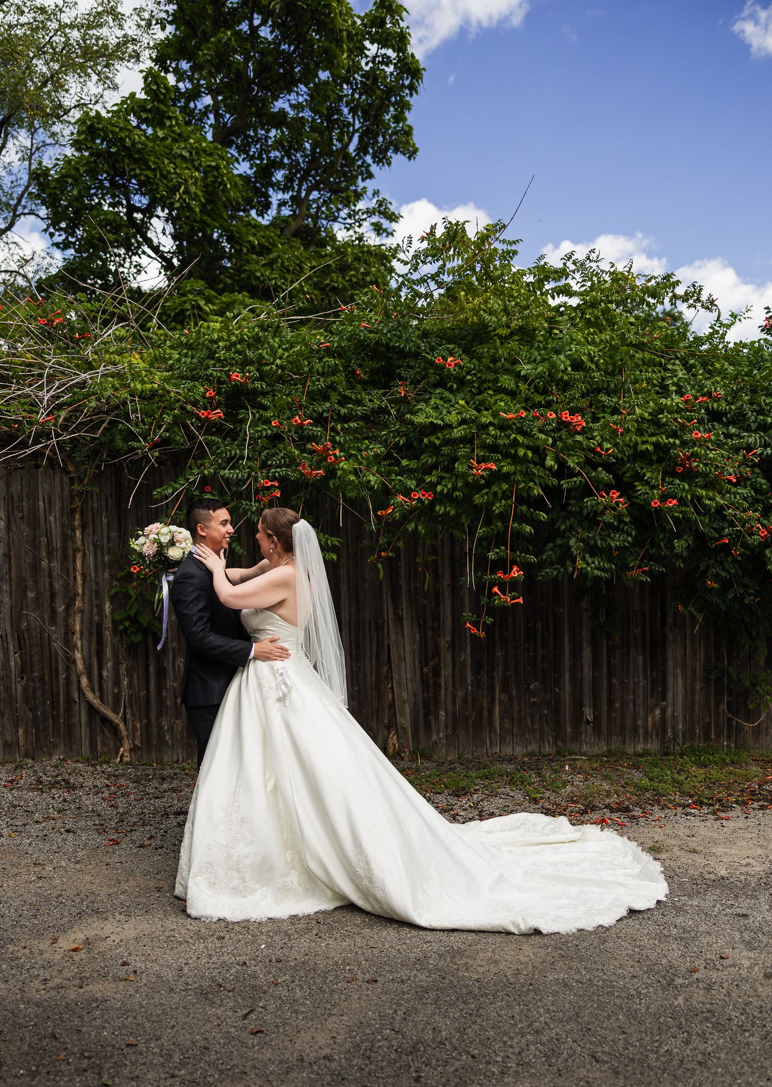 Bride and groom embracing in front of a wooden fence with lush green foliage.