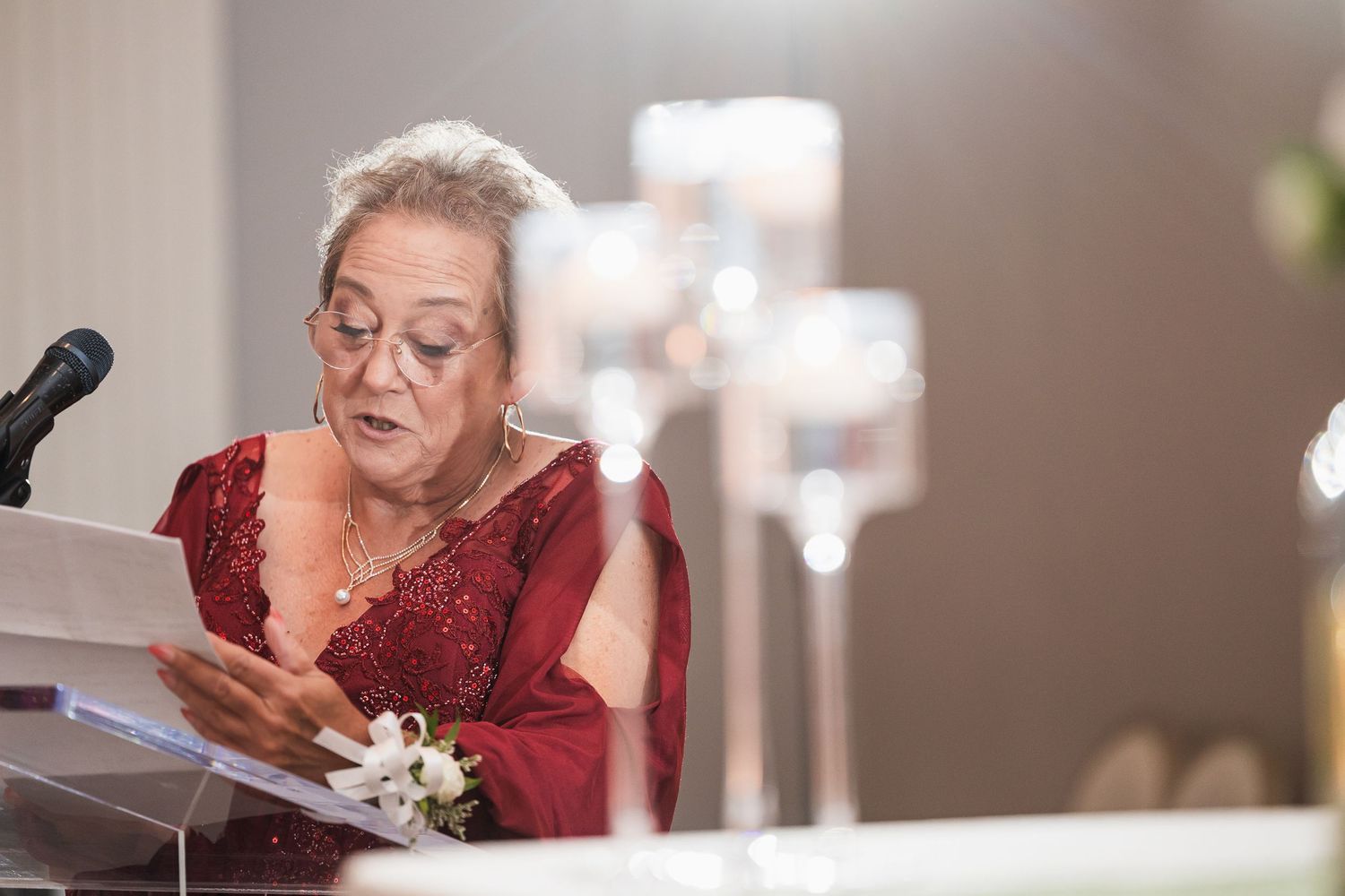 Elderly woman in red dress reading speech at wedding reception podium with candles in background.