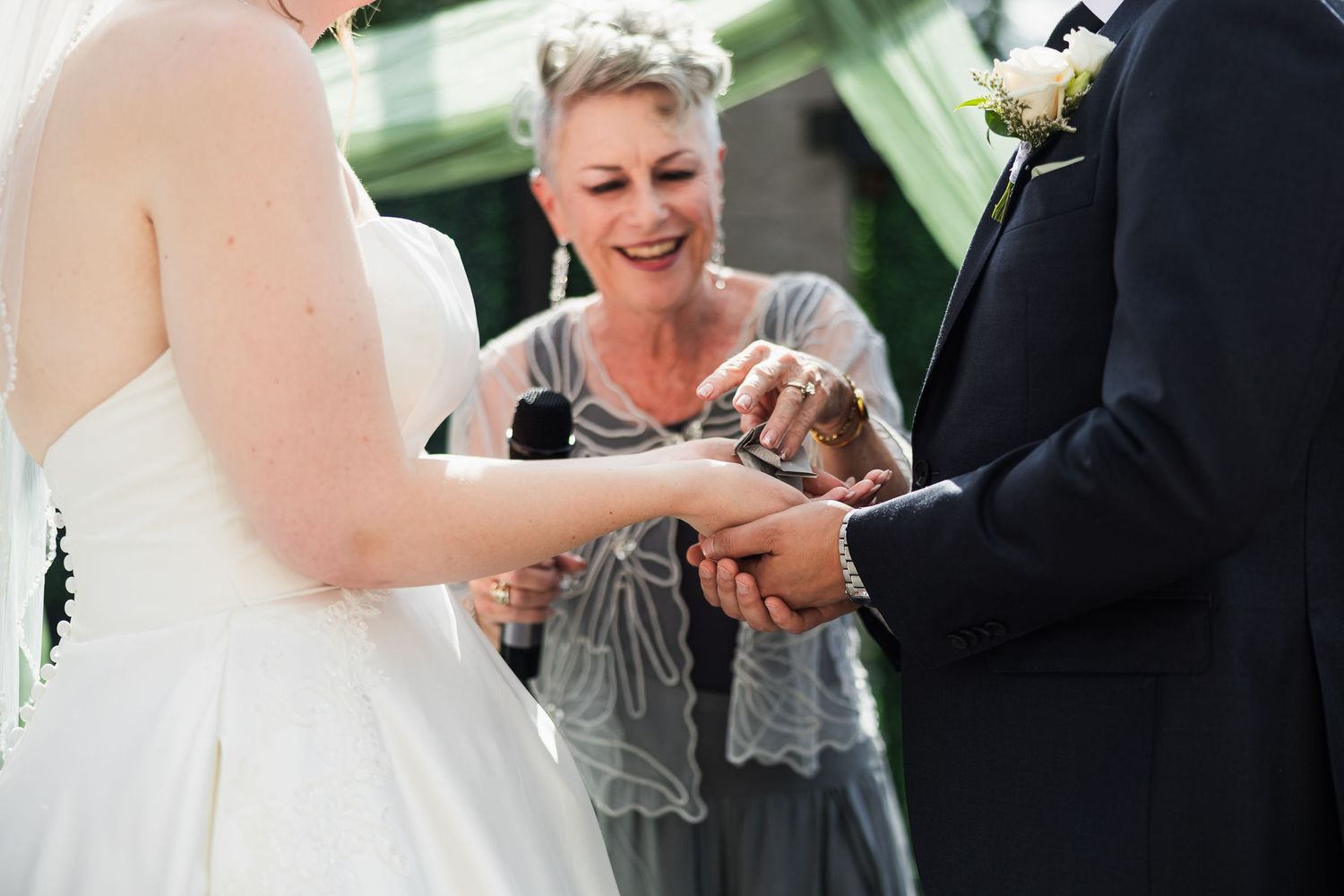 Officiant placing wedding rings on bride and groom hands during outdoor wedding ceremony.