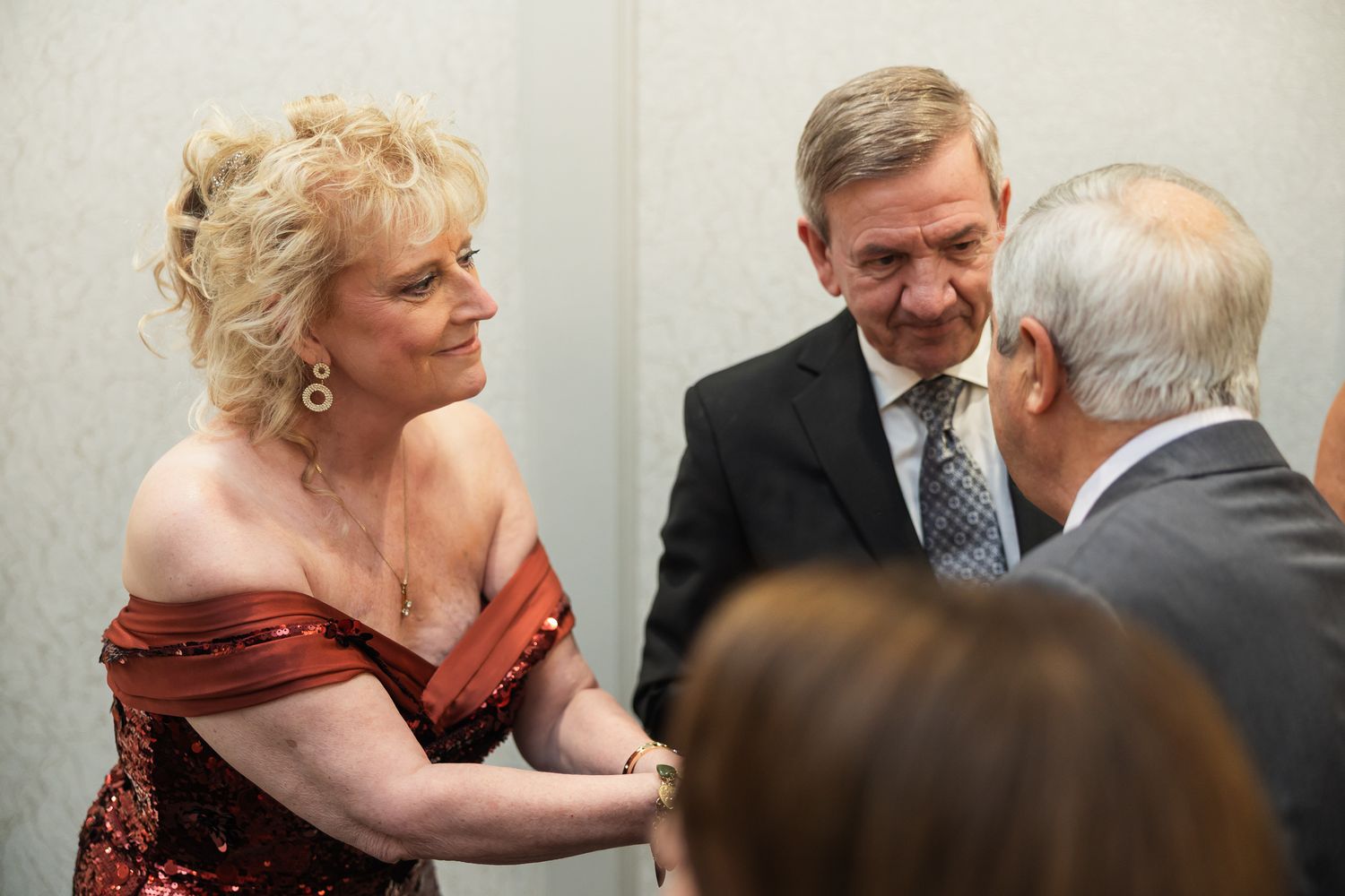 Elegantly dressed woman in red sequin gown conversing with two men in suits at a formal event.