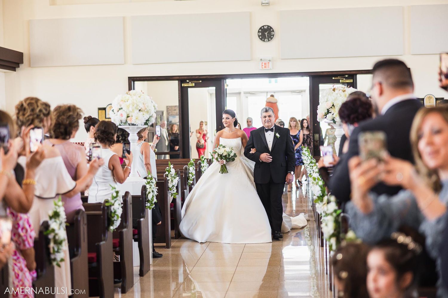 Wedding ceremony processional in a church with guests taking photos from the pews.