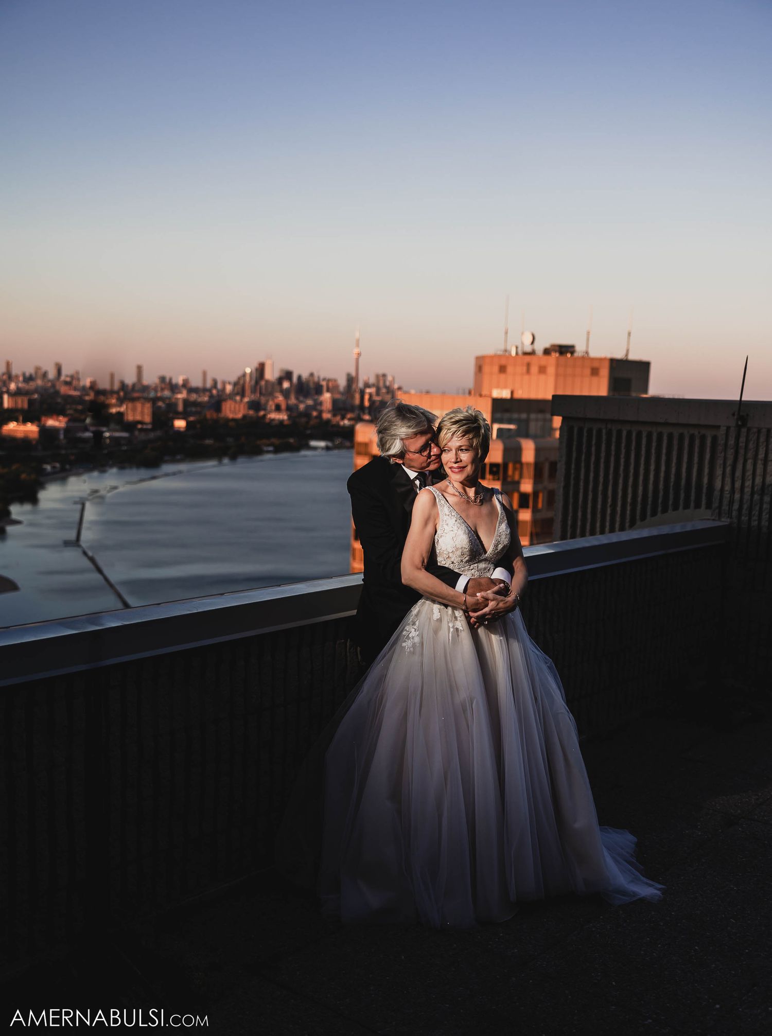 A bride in a white ballgown stands with companion against a sunset city skyline.
