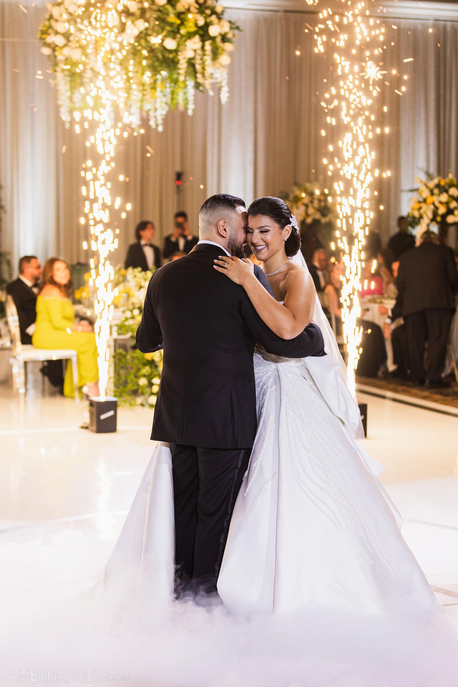 Couple dances beneath sparkling lights and floral arrangements with fog effects on dance floor.