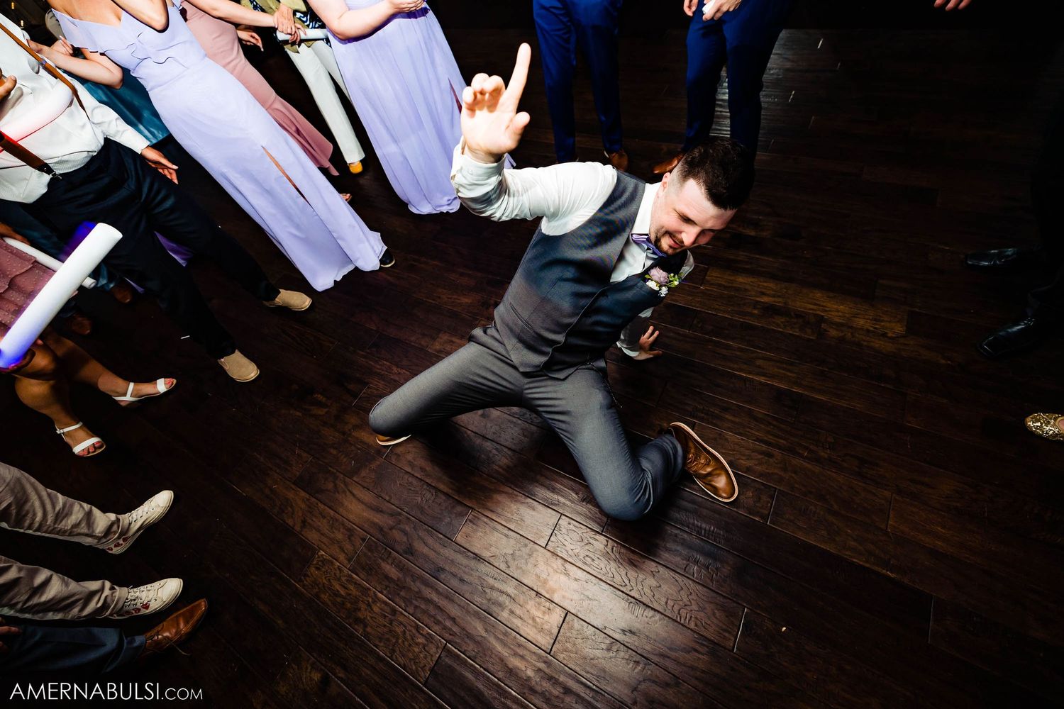 Wedding guest performing acrobatic dance move on wooden floor surrounded by partygoers.