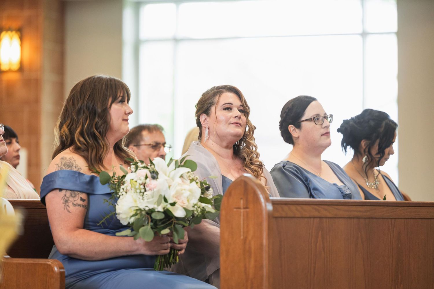 Bridesmaids in blue dresses holding white floral bouquets seated in church pews.
