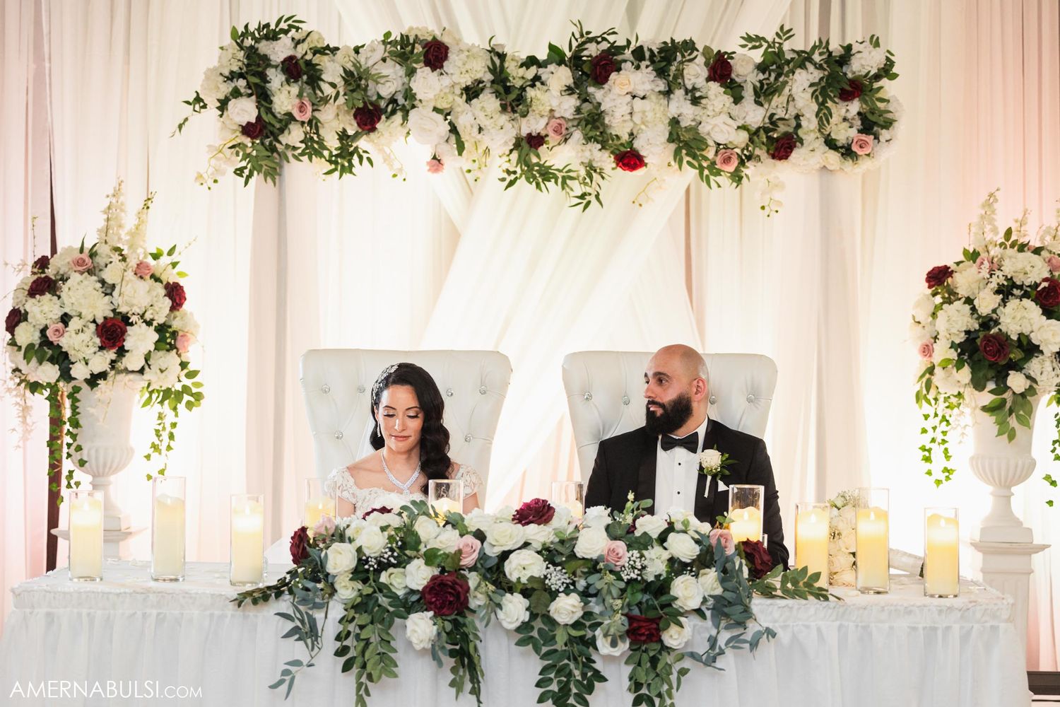 Newlyweds seated at decorated sweetheart table with floral arch and elegant white decor.