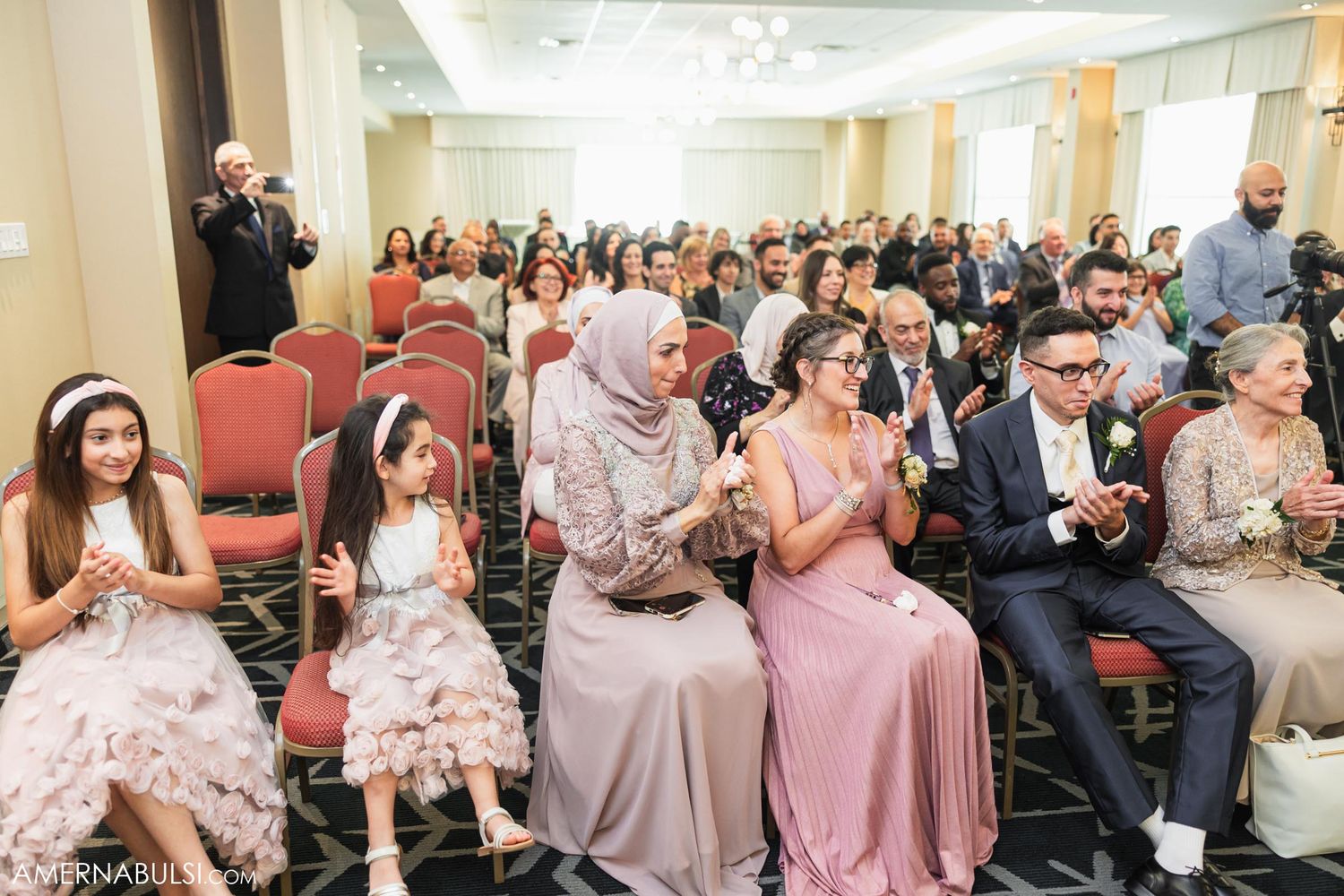 Wedding guests in pastel dresses seated in rows during an indoor ceremony.