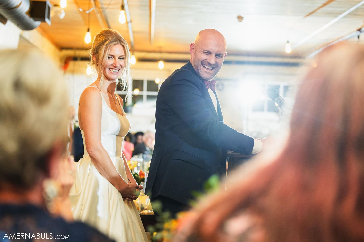 A couple laughs while giving their wedding speech in a rustic reception venue.