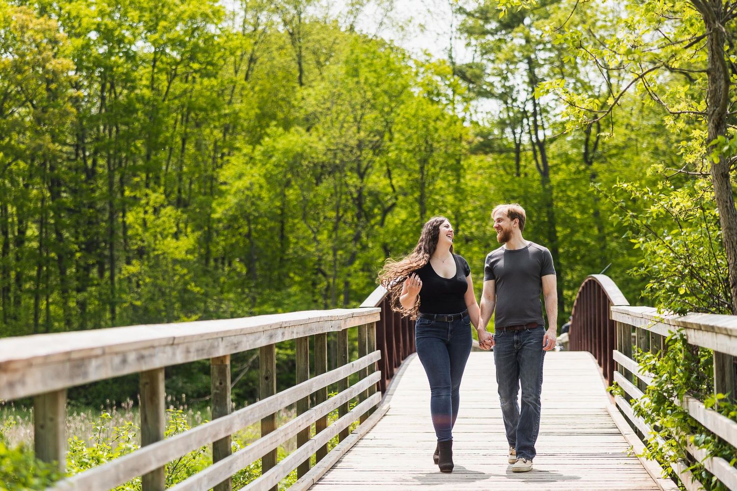 Couple walking hand in hand on wooden bridge surrounded by green forest.