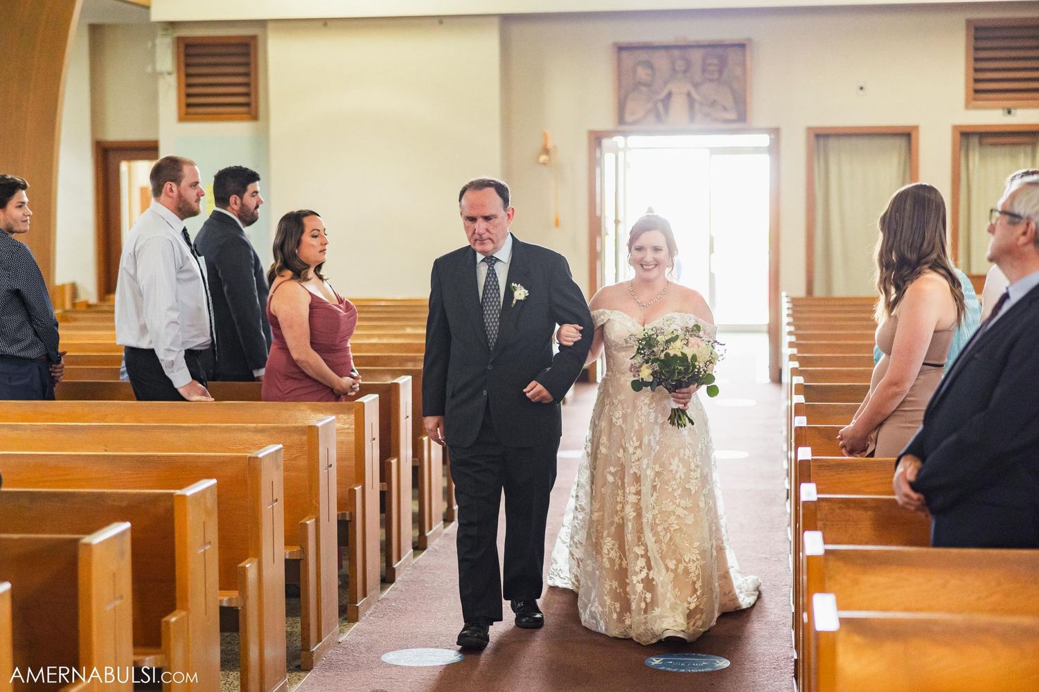 A wedding ceremony procession moves down the aisle of a church with wooden pews and natural lighting.