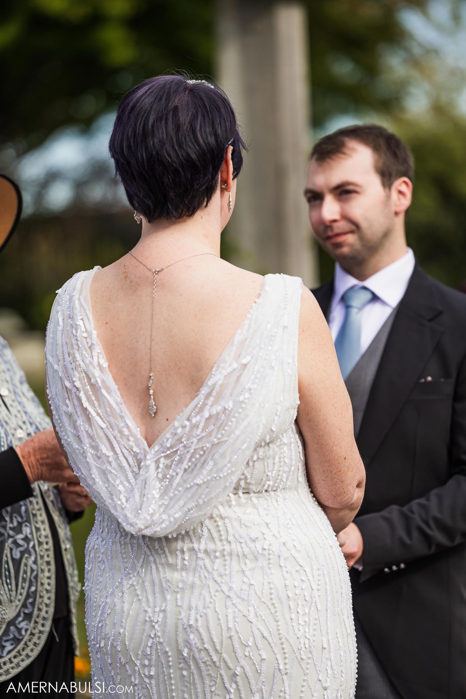 Back view of a bride in an elegant white lace dress with a plunging V-back at an outdoor ceremony.
