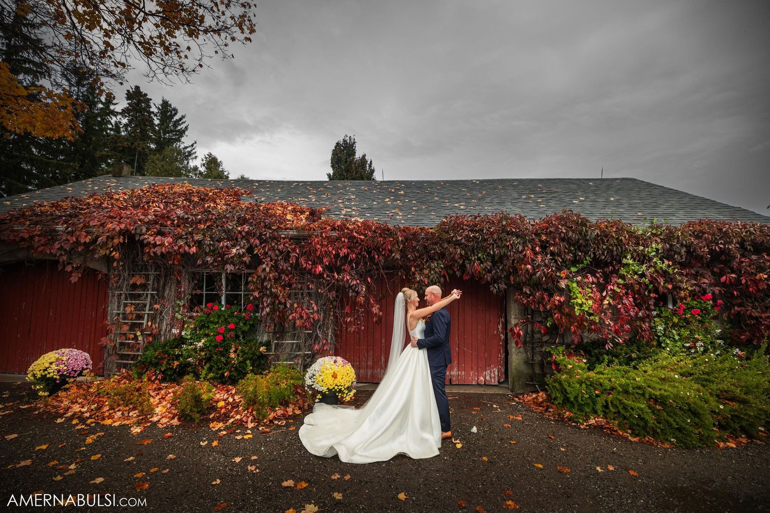 Newlyweds pose against red barn with autumn foliage and fall decorations.