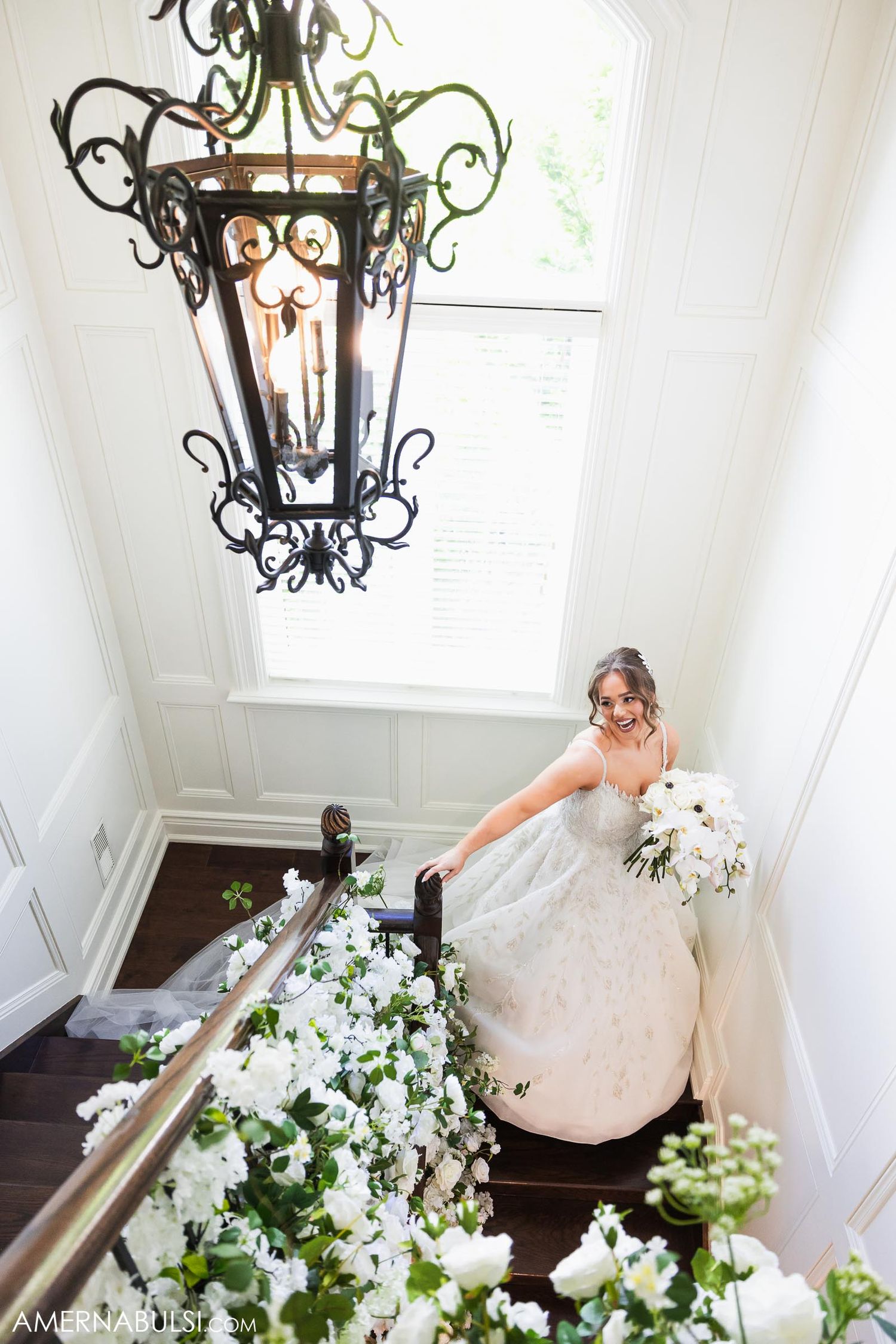 A dramatic wrought iron chandelier hangs above a staircase adorned with white floral arrangements.